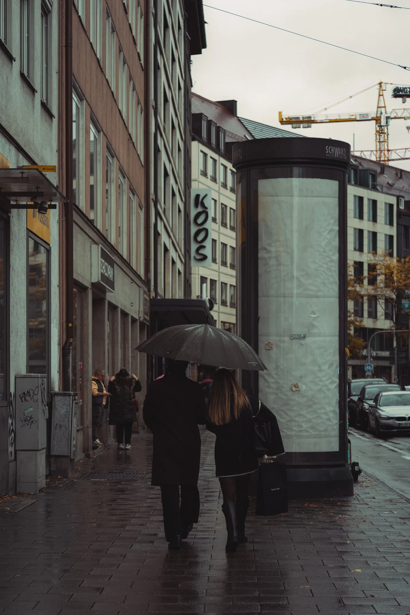 People walking on a rainy city street with umbrellas, surrounding buildings, and construction cranes in the background.