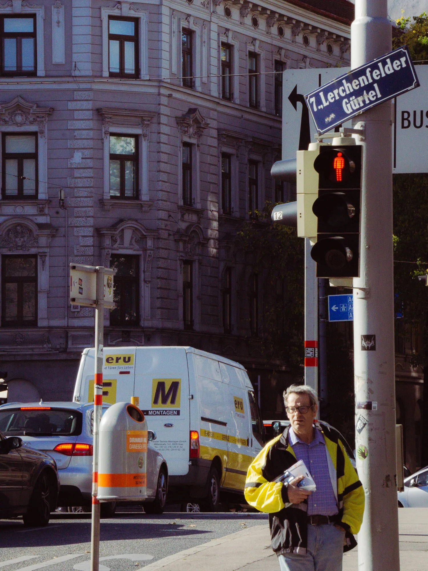 City street scene with a man wearing a yellow jacket holding papers, standing near a pole with street signs. Behind him, there are several vehicles including a white van with yellow and purple signage, and a dark-colored car. An ornate building with 