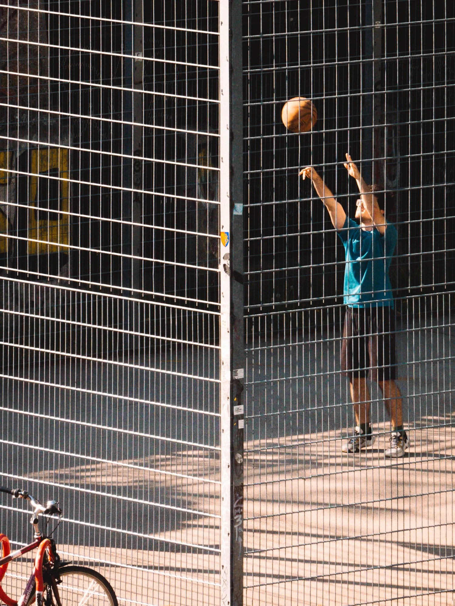 A person wearing a teal shirt and dark shorts is playing basketball behind a metal fence at a basketball court. The individual appears to be shooting the ball towards the hoop. The court is outdoors with a dark, possibly black, wall in the background
