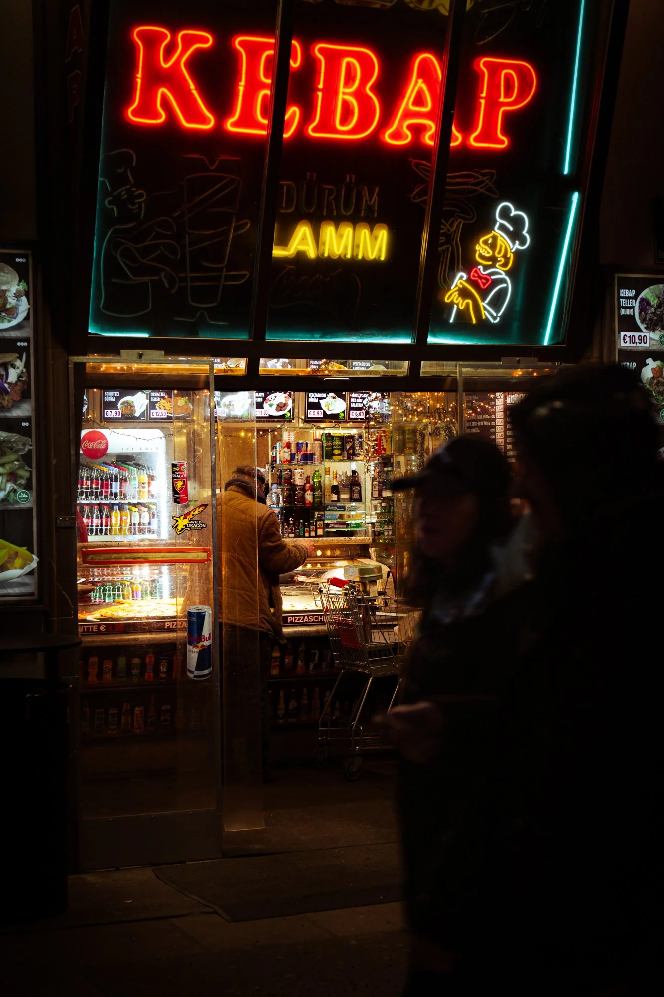Neon sign outside a kebab shop that reads 'KEBAP' in large red letters, with smaller signs for 'DÜZÜM' and 'AMM'. The shop has a lit interior with a glass display case showing food items, and a person wearing a brown jacket standing behind it. There 