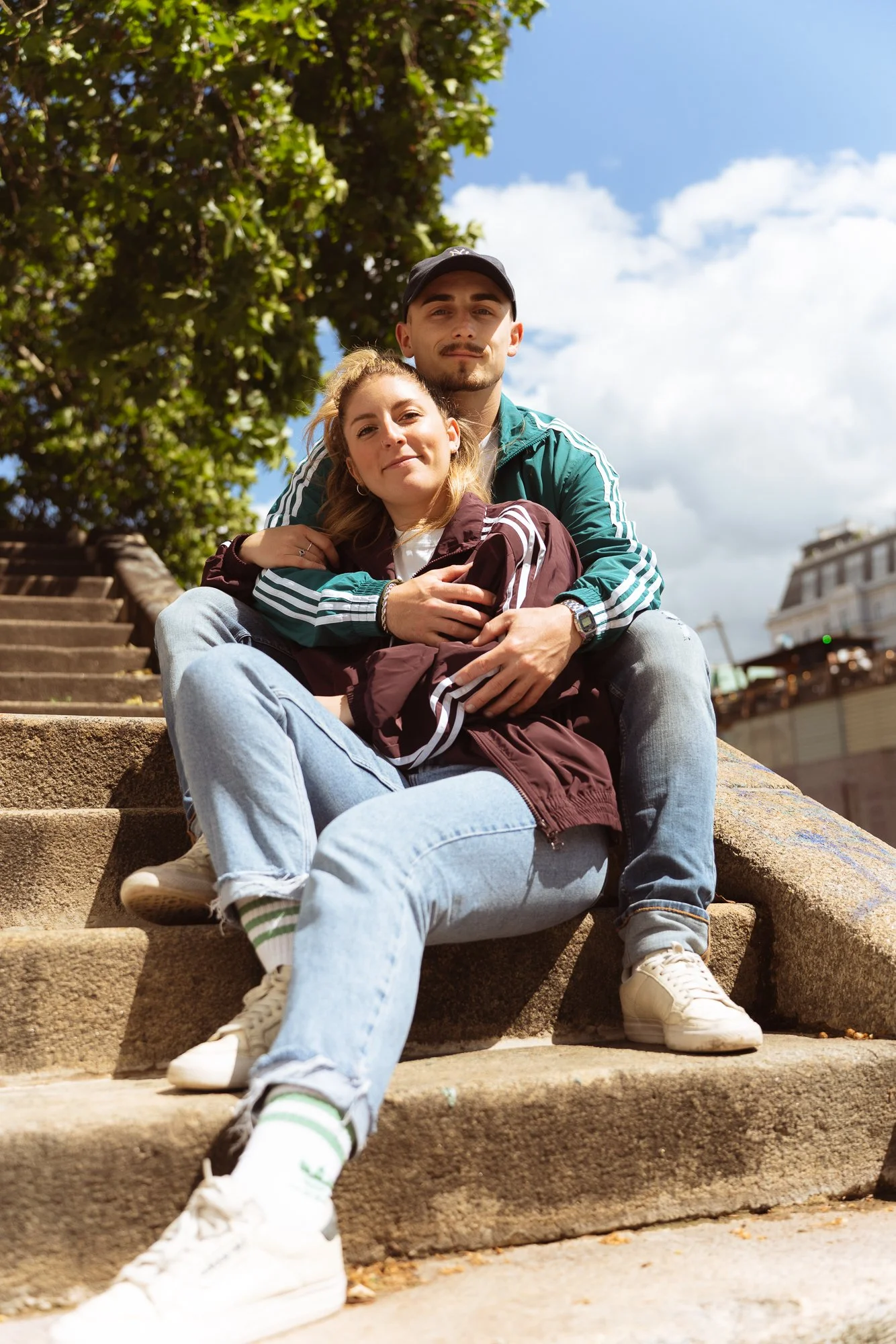 A young man and woman sitting on outdoor steps, with the woman leaning against the man. They are smiling, and the background features a large tree with green leaves, a blue sky with clouds, and buildings.