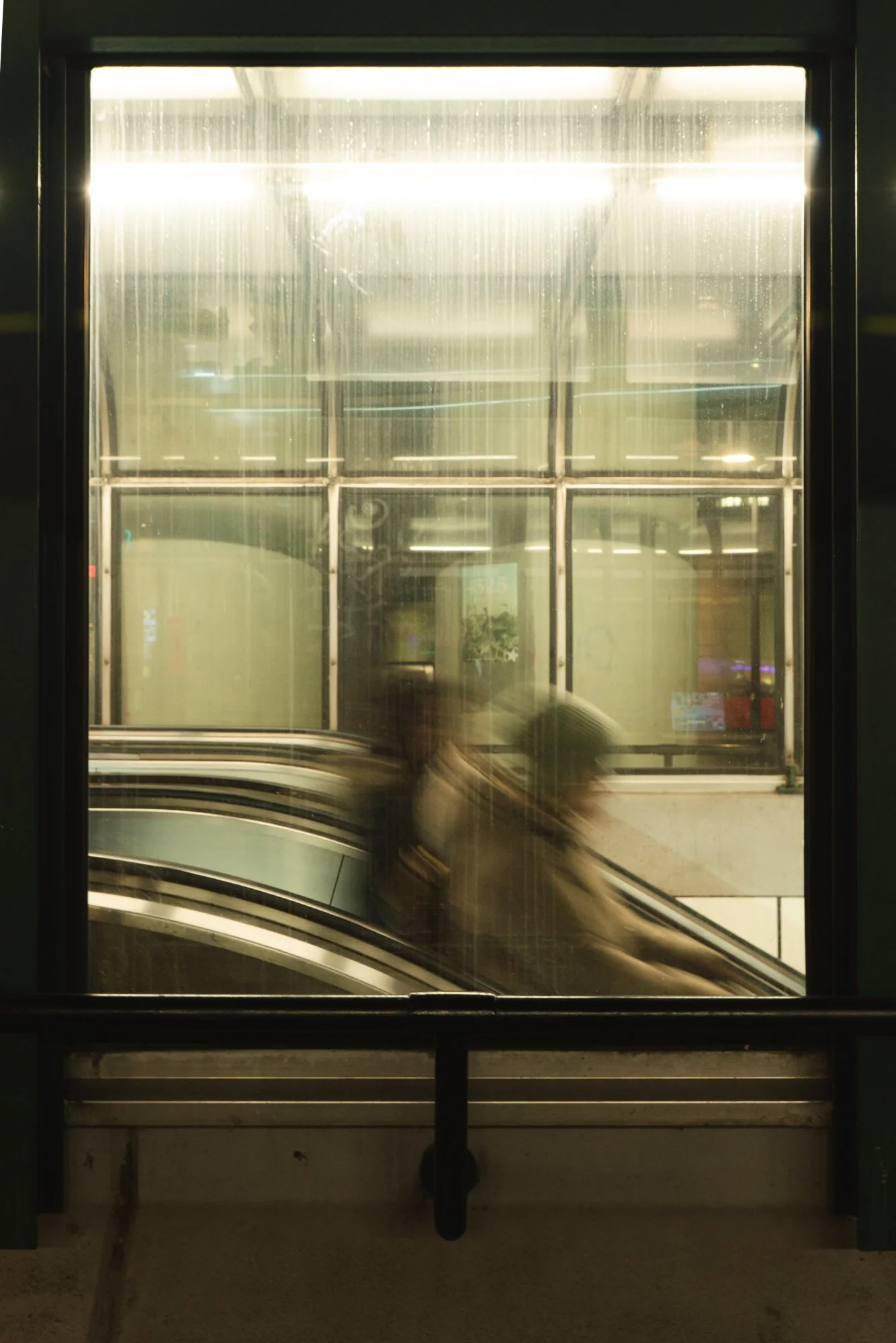 View of a large glass window with reflections and a blurry escalator inside a building, possibly an airport or shopping mall, with bright overhead lighting and a person partially visible.