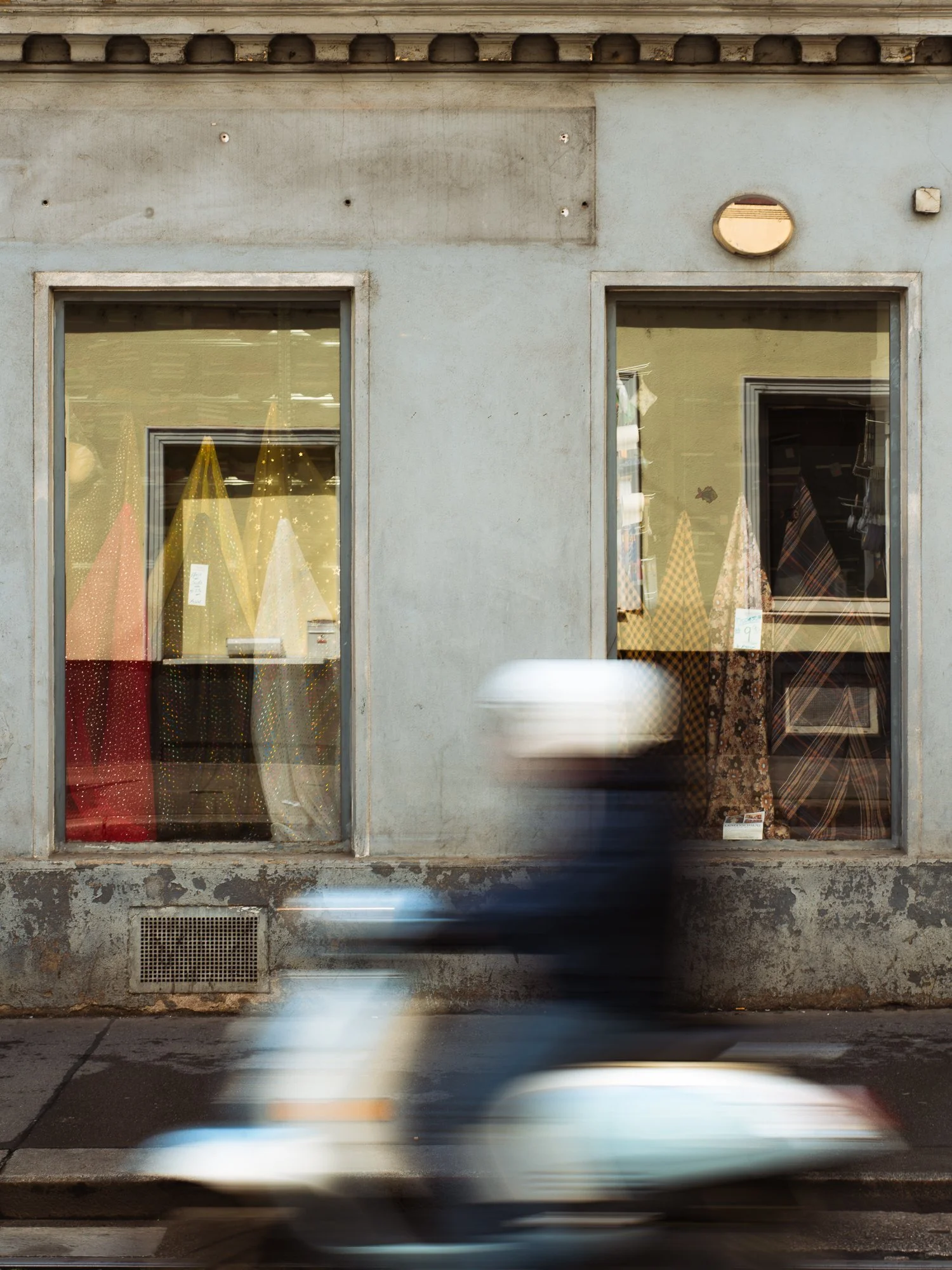 Storefront with two large windows displaying fabric or textile items, with a blurred vehicle passing in front.