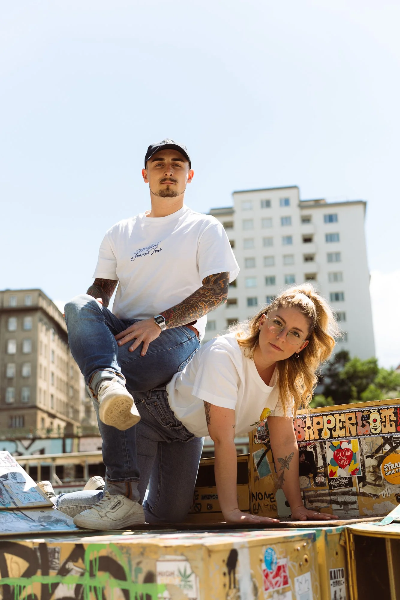 Two individuals, a man and a woman, are posing creatively on a city street in front of a graffiti-covered object, with buildings in the background. The man is crouching on one knee with one arm resting on his leg, wearing a white t-shirt, jeans, and 