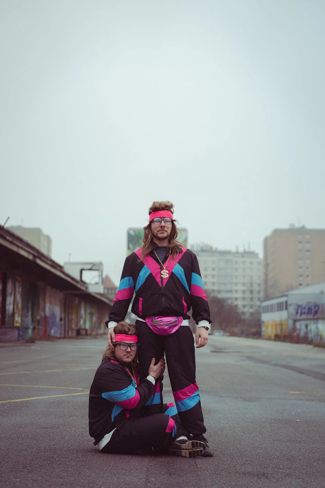 Three men dressed in 80s style workout clothes with bright colors, long hair, and glasses, posing with one standing and two kneeling on an empty urban street with buildings in the background.