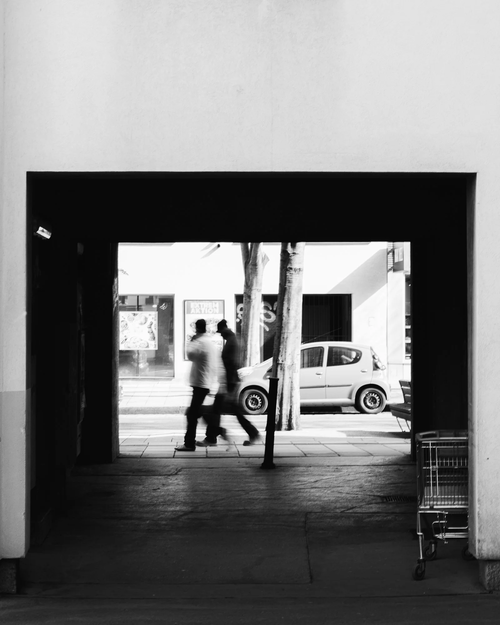 Black and white photo taken from inside an opening, looking out onto the street with a sidewalk, trees, and a parked car. Two people are walking past each other across the street, and there are shop windows and signs in the background.