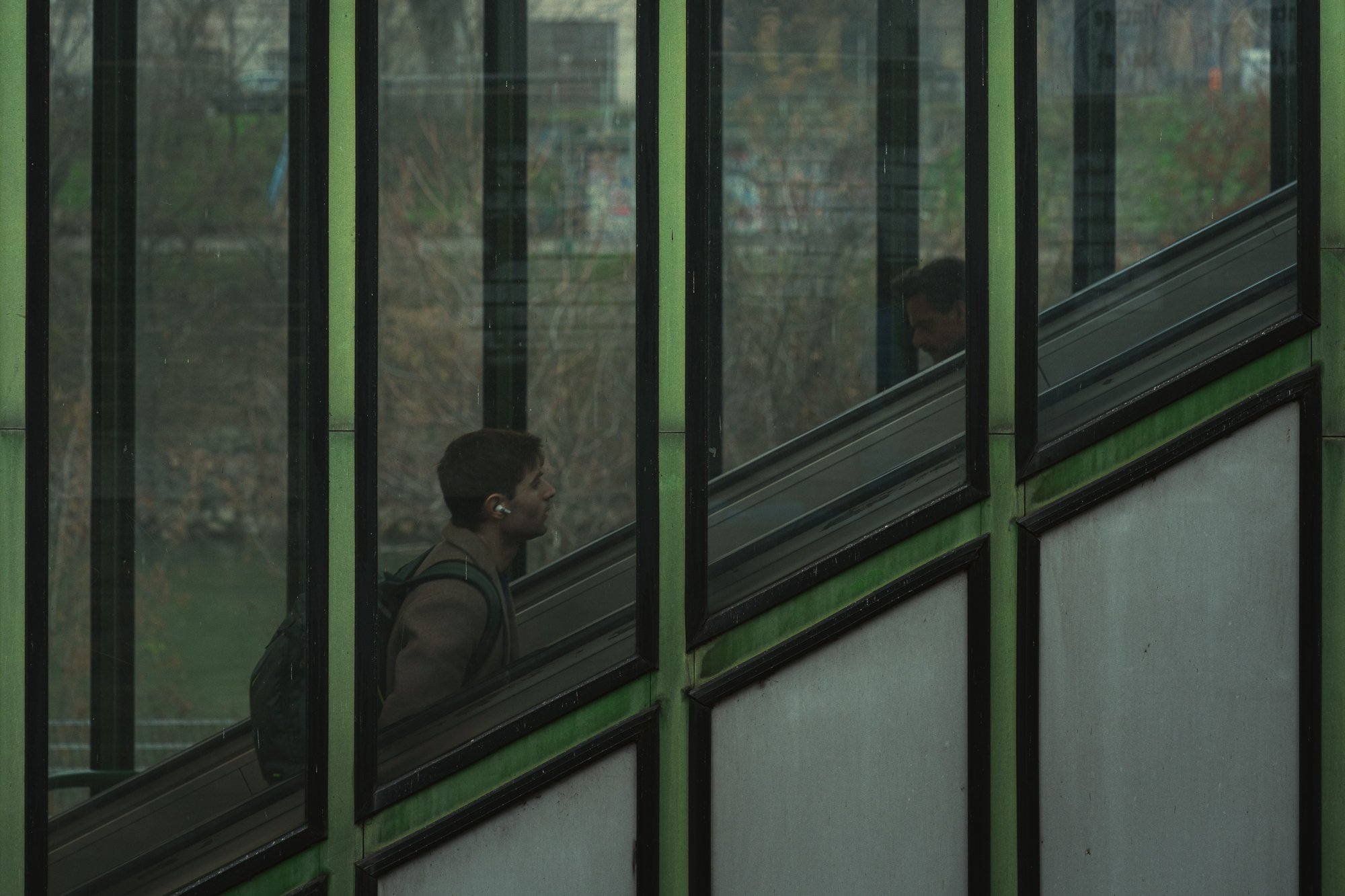 A person with a backpack walking past a glass window with green framing, seen from inside a building.