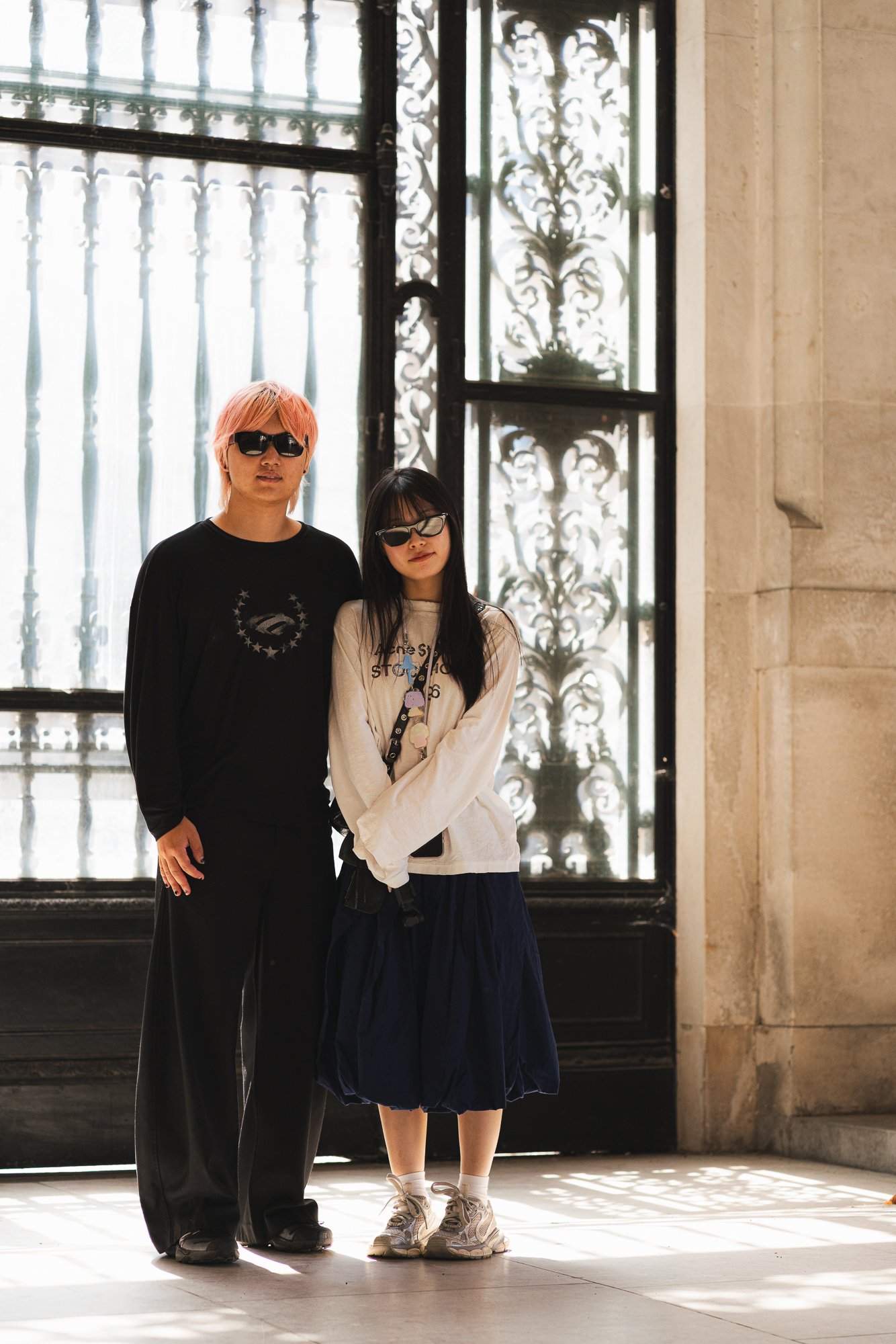 Two young women with sunglasses standing in front of a decorative metal and glass door, with sunlight streaming through. The woman on the left has pink hair and is wearing a black shirt, while the woman on the right has black hair and is wearing a li