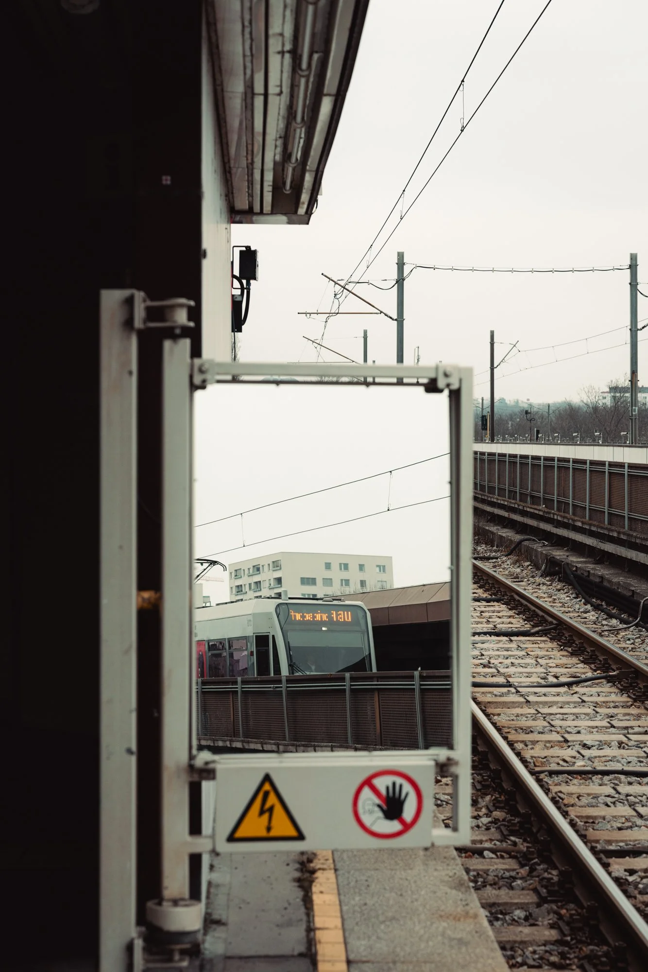 View of a train station platform with a train approaching, overhead electrical wires, and safety barriers with warning signs.