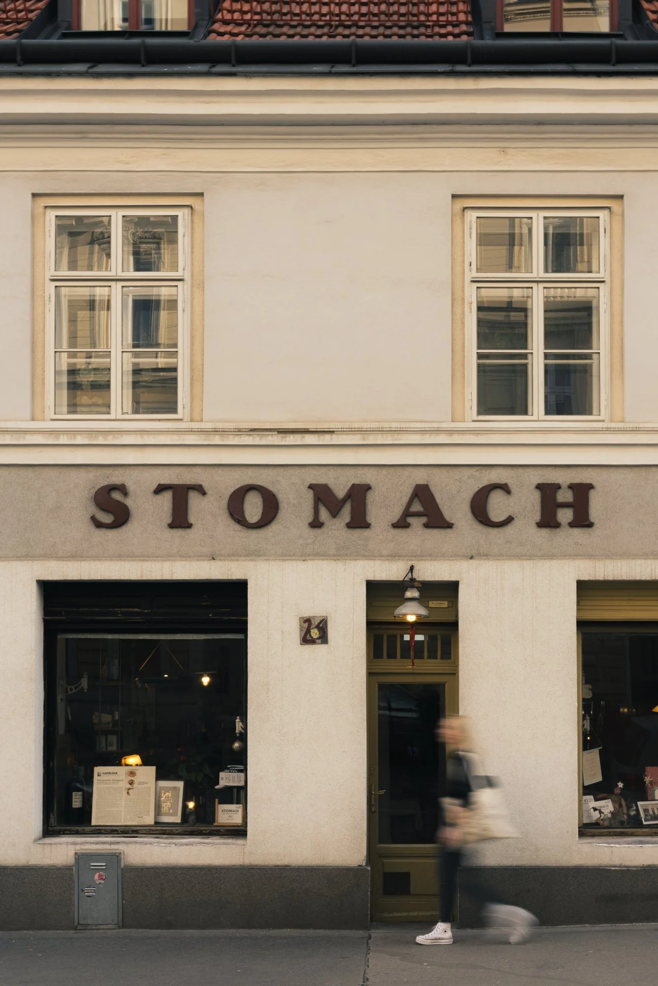 The facade of a building with two large windows and a door in the center. The building has a sign that reads 'STOMACH'. A person wearing white sneakers, dark pants, and a jacket is walking past, appearing blurred due to motion.