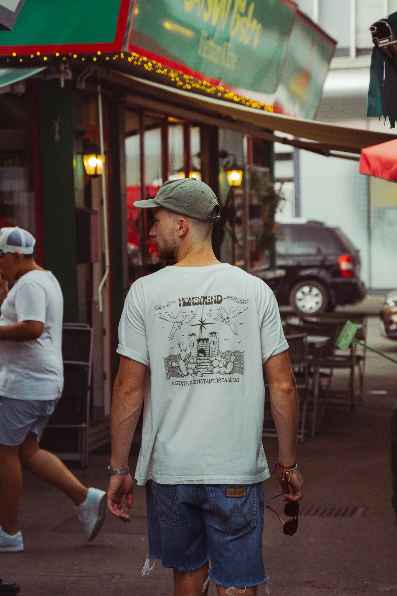 A man wearing a light gray cap and a white t-shirt with a graphic design and text on the back, standing outdoors near a market or street food stall. The background shows a black vehicle, some tables, and a person in a white shirt and cap.