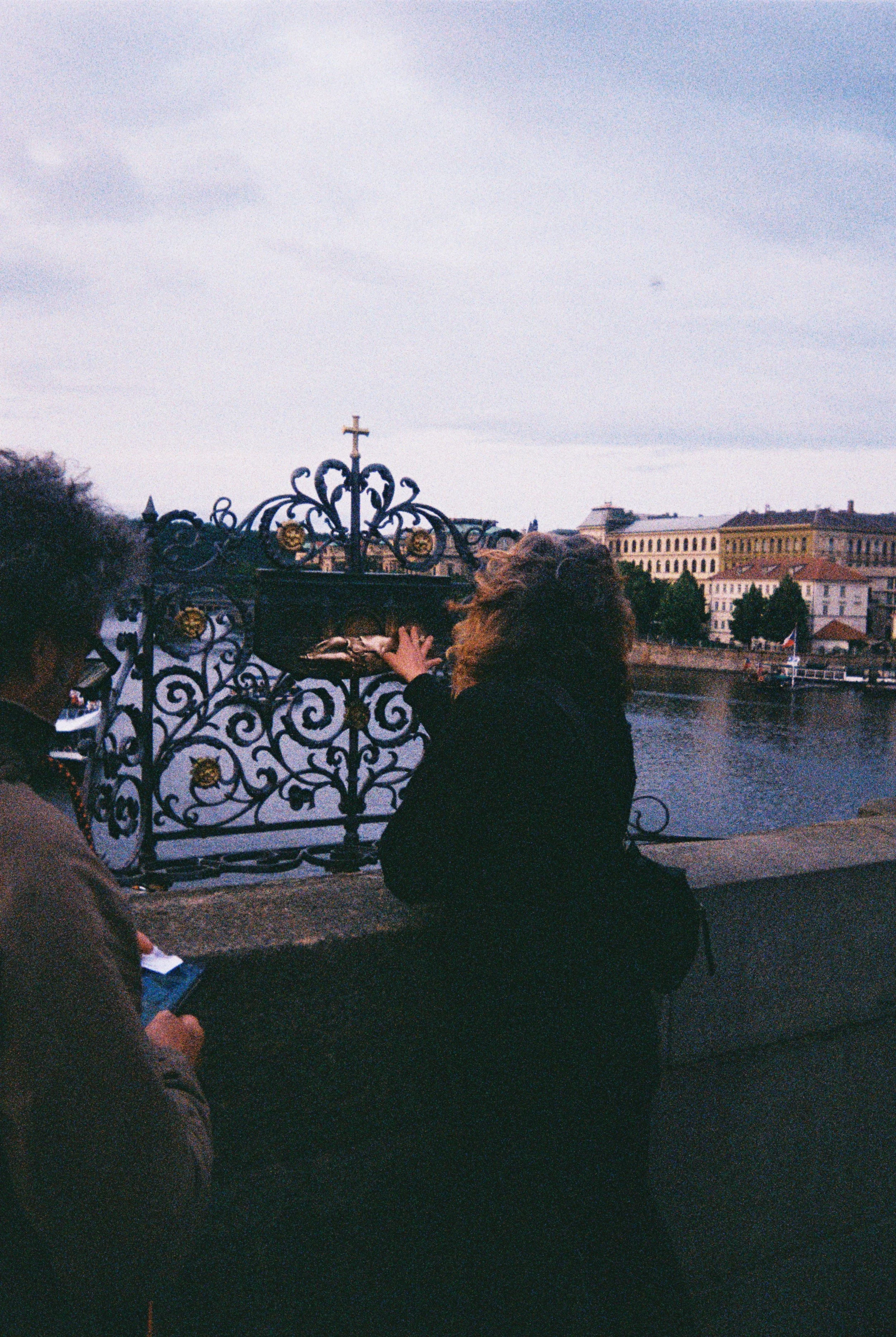 People sitting by a river, with a black wrought-iron gate featuring gold and a cross in the foreground, and historic buildings in the background under cloudy skies.