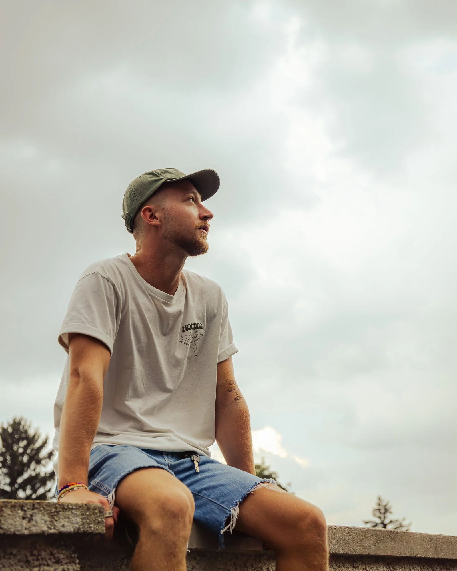 A young man with a beard and mustache wearing a gray baseball cap and a white T-shirt with a graphic design sits outdoors on a wooden surface, looking to the right. The background shows a cloudy sky and trees.