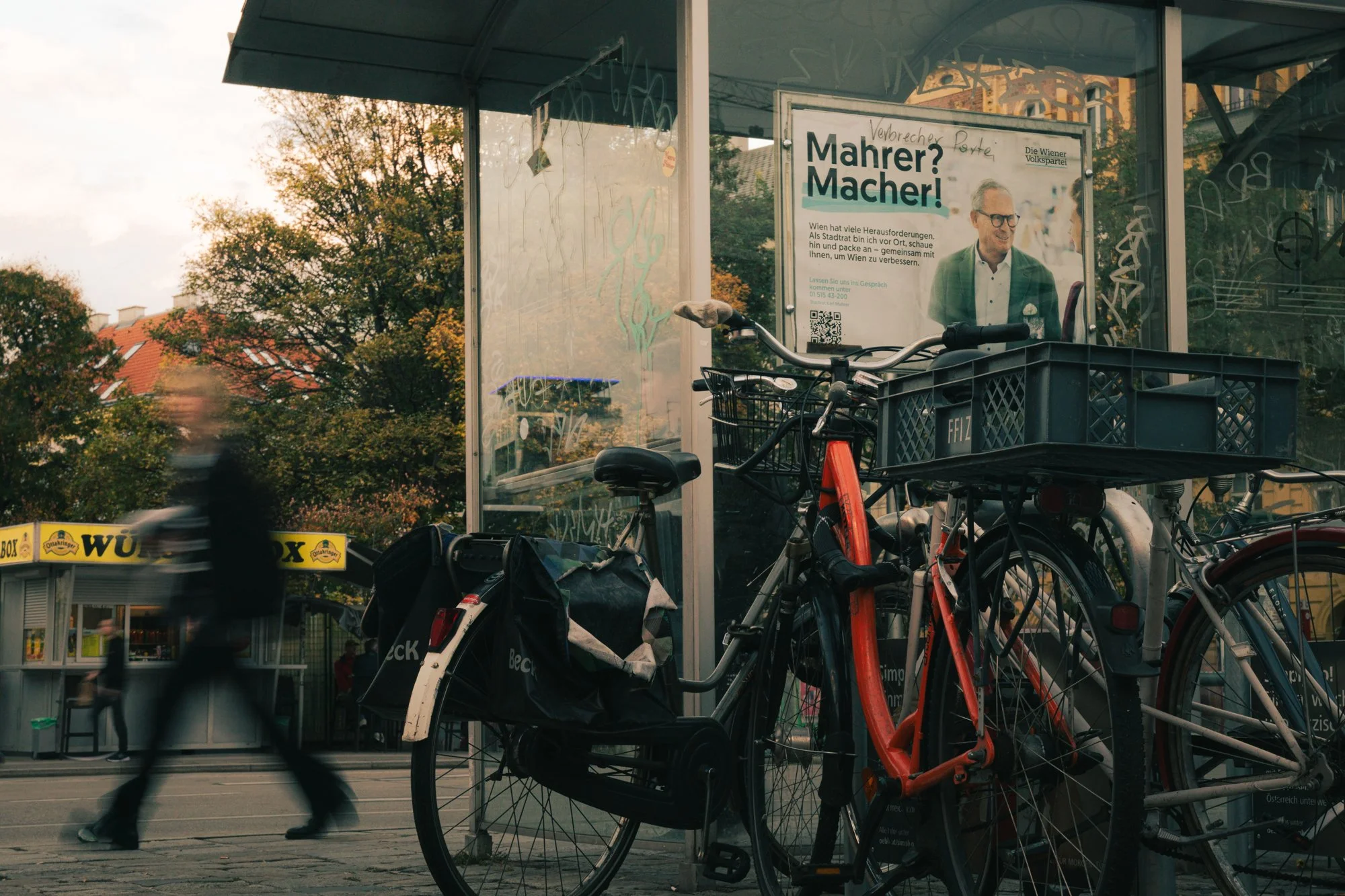 A bus stop shelter with bicycles parked in front, a blurred pedestrian walking past, and a billboard inside the shelter with a smiling man in glasses and a green jacket, and text in German.