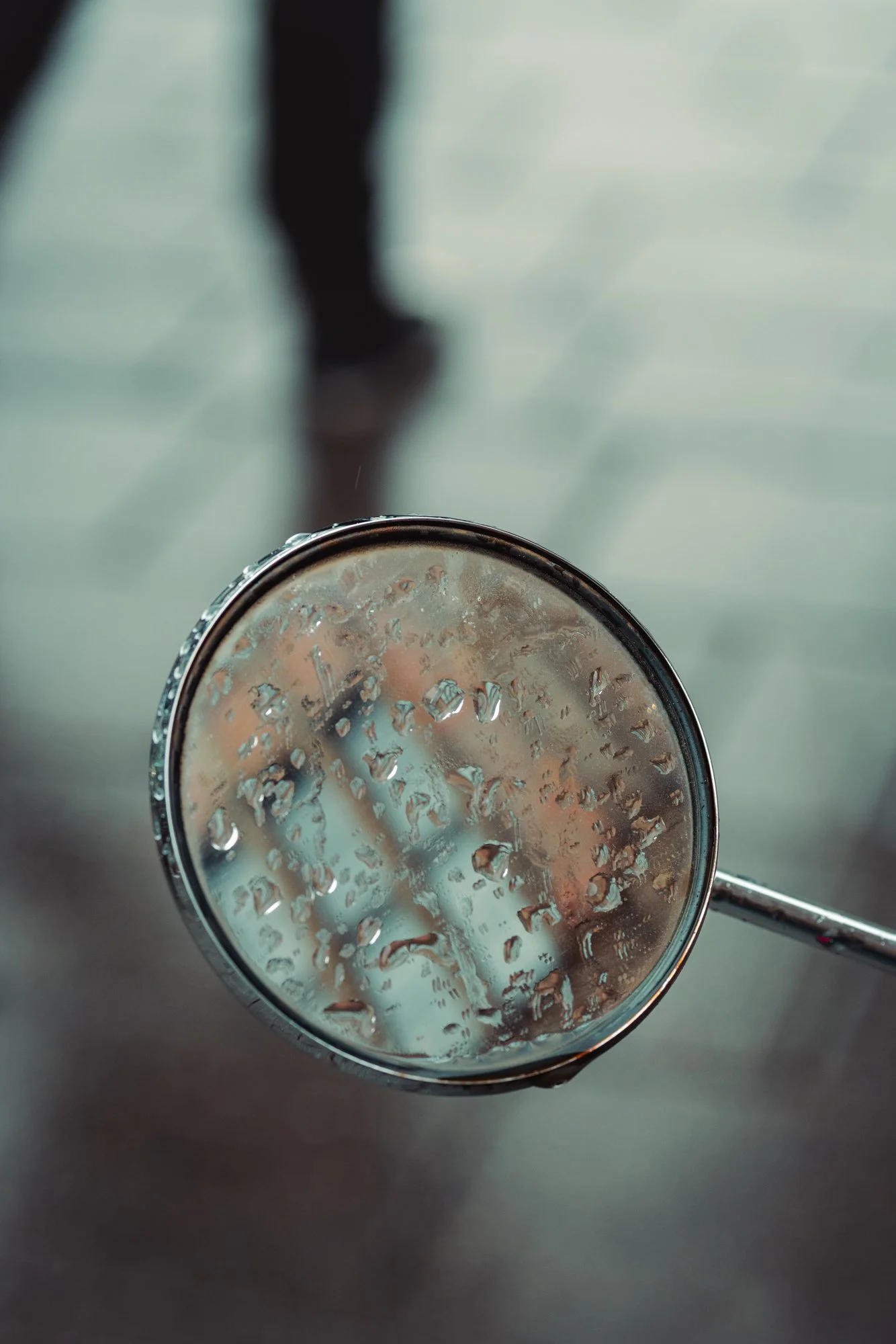 Close-up of a stainless steel spoon with water droplets on it, held over a reflective surface, with blurred human legs in the background.