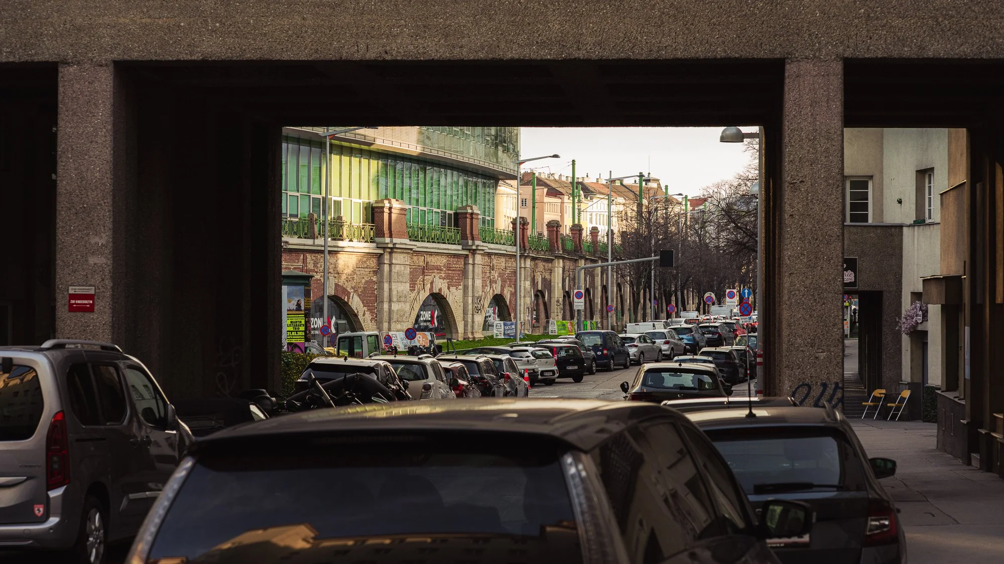 City street viewed through a large archway, with parked cars along the street and a modern building with green glass windows in the background.
