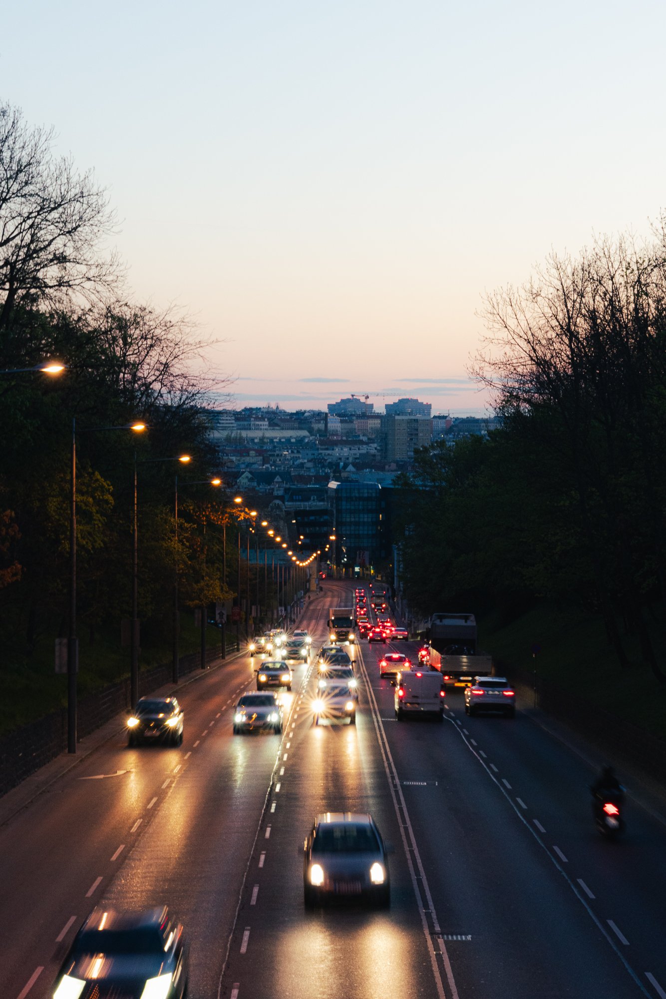 City street during evening rush hour with cars driving, street lamps on, and city skyline in the background with buildings and trees.