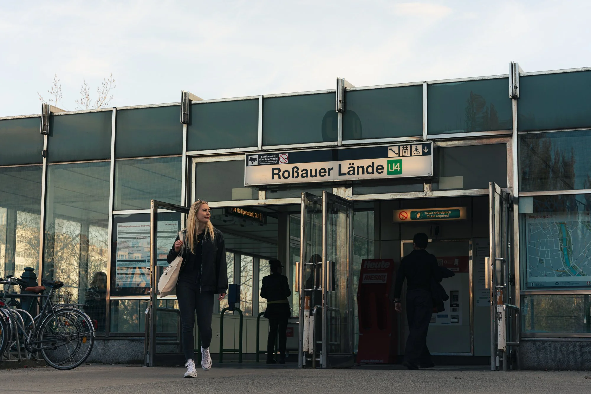People entering and leaving the Roßauer Lände U4 subway station in Berlin, with bicycles parked outside and a woman walking out of the station.
