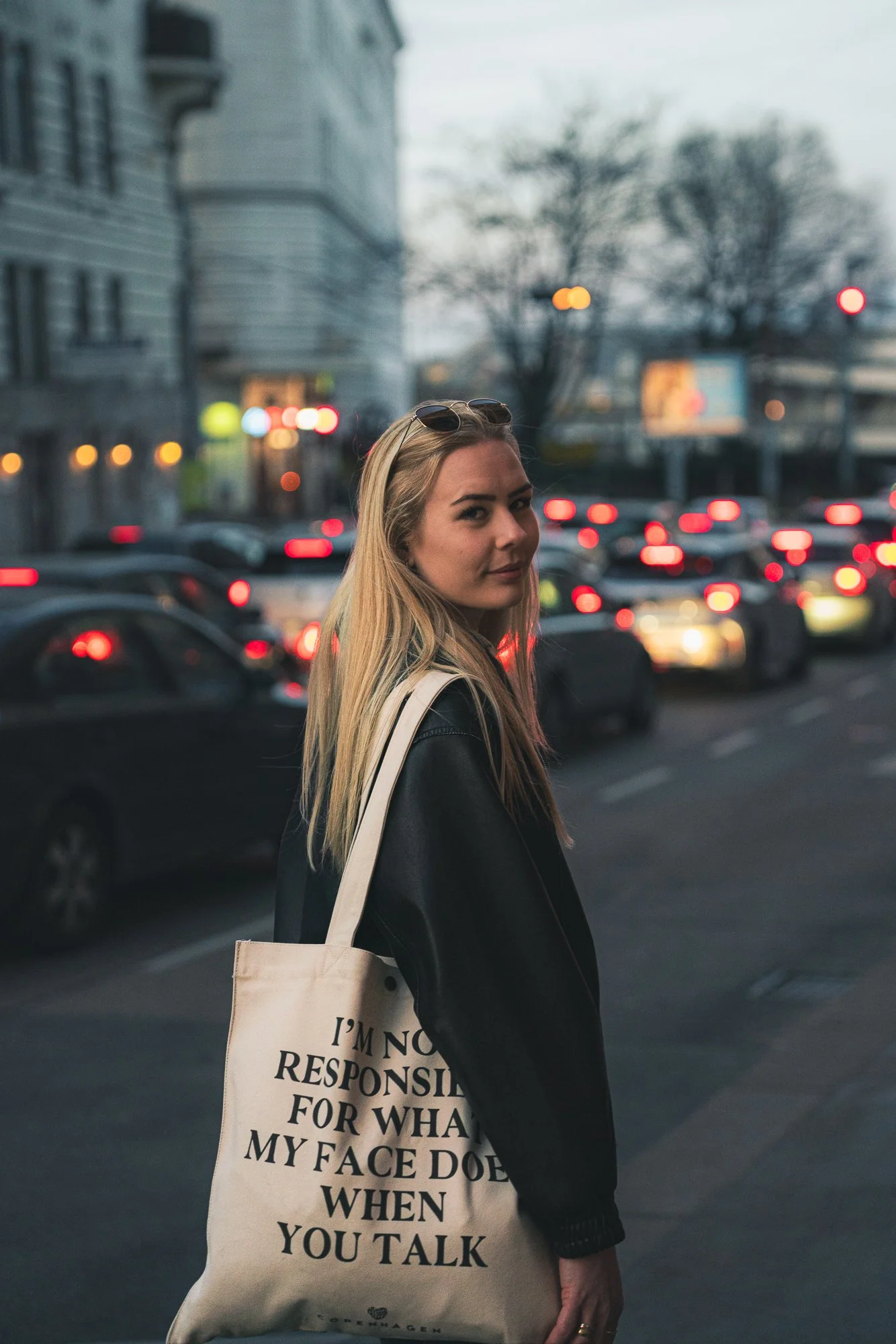 A young woman with blonde hair, wearing dark sunglasses on her head, dressed in a black jacket, carrying a large beige tote bag with black text. She is standing on a city street with cars and blurred city buildings and trees in the background during evening or dusk.