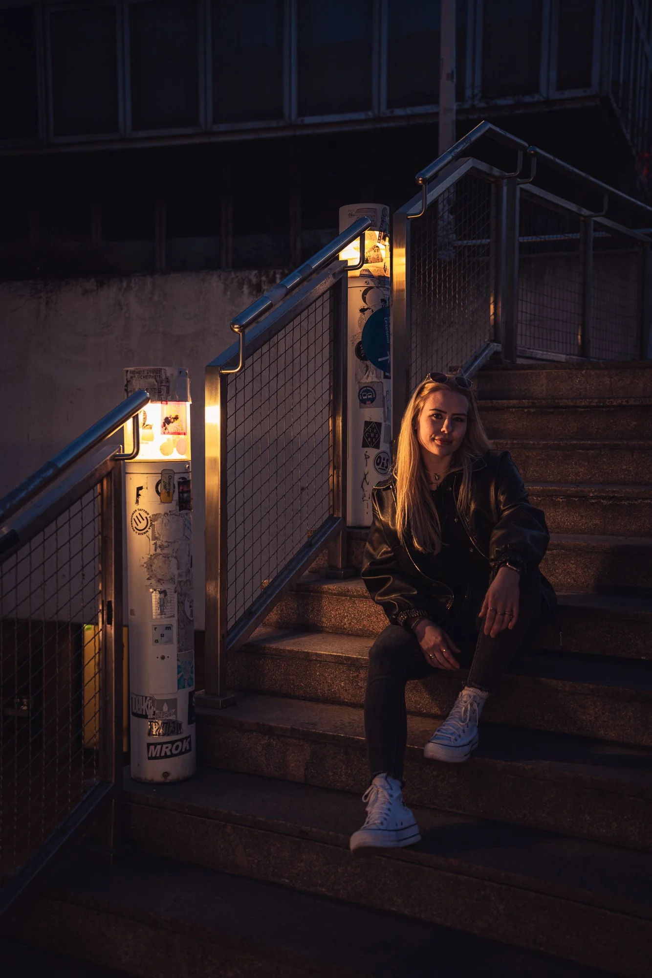 A young woman with blonde hair sitting on outdoor stairs at dusk, wearing a black leather jacket, jeans, and white sneakers, with sunglasses on her head. The stairs are illuminated by warm lights, and there are graffiti-covered pillars nearby.