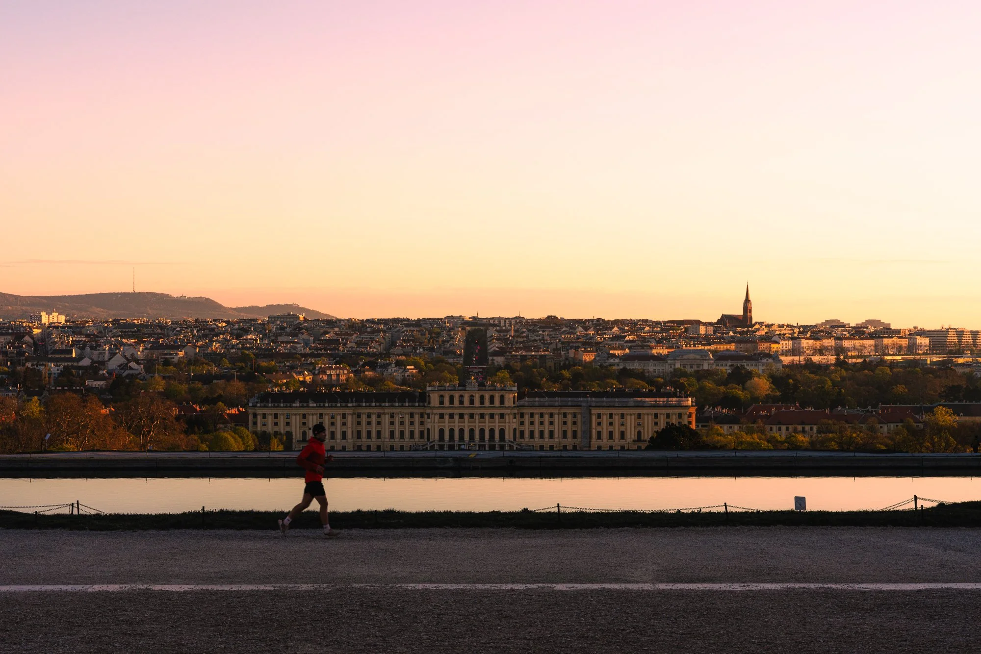 A person jogging along a riverwalk at sunset with a city skyline and distant hills in the background.