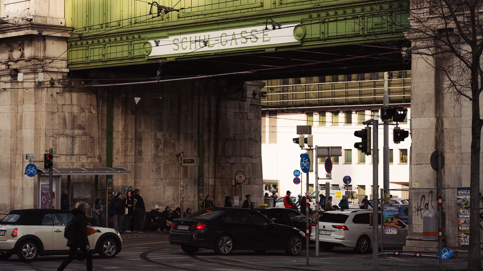 City street scene under a bridge with people waiting at a bus stop, cars, bicycles, traffic lights, and signs. The bridge has a sign that reads 'Schul-Gasse'.