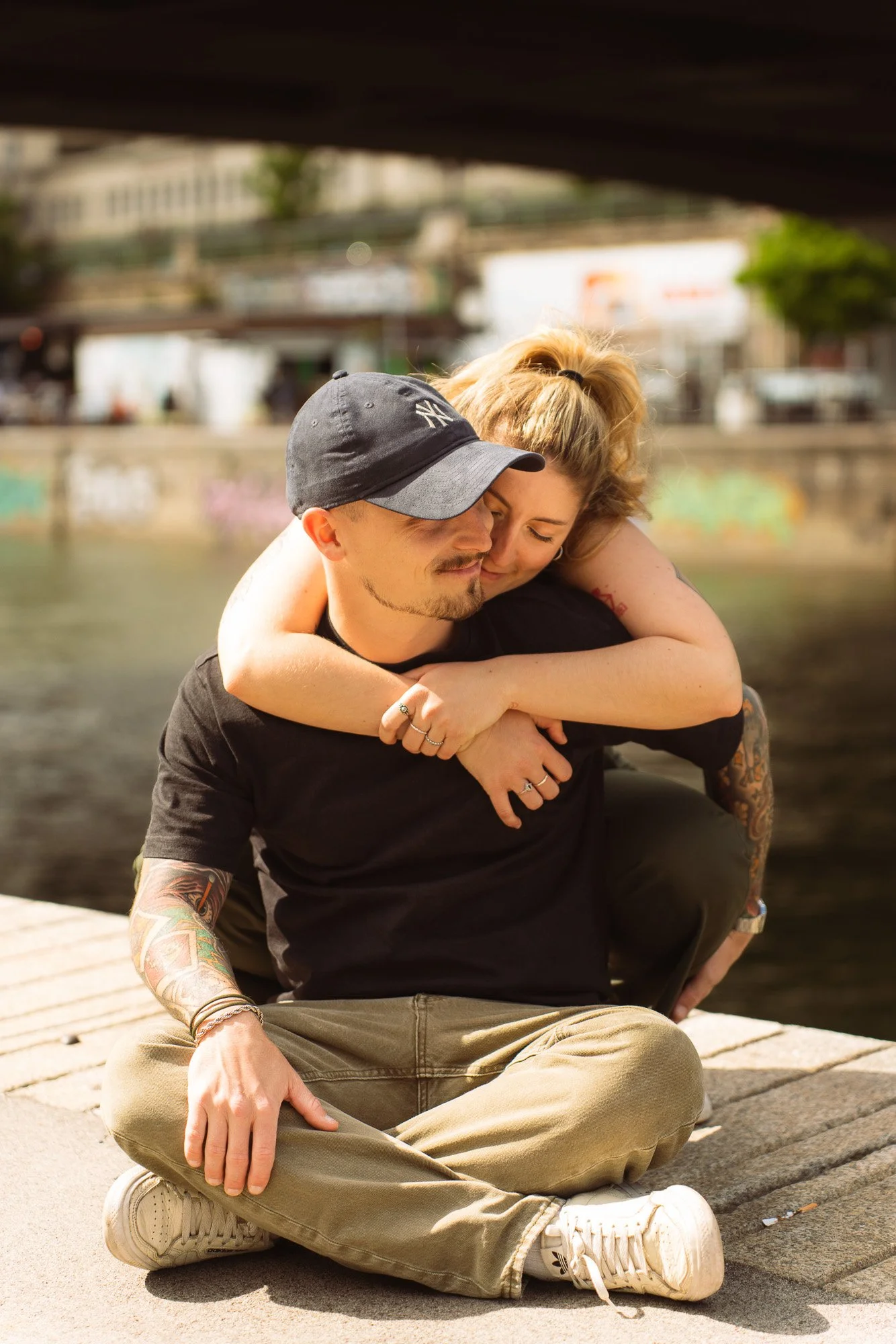 A man and woman sitting outdoors on a dock, embracing each other with closed eyes. The woman has blonde hair tied in a ponytail, and the man is wearing a black baseball cap. The background features a river with blurred buildings and trees.