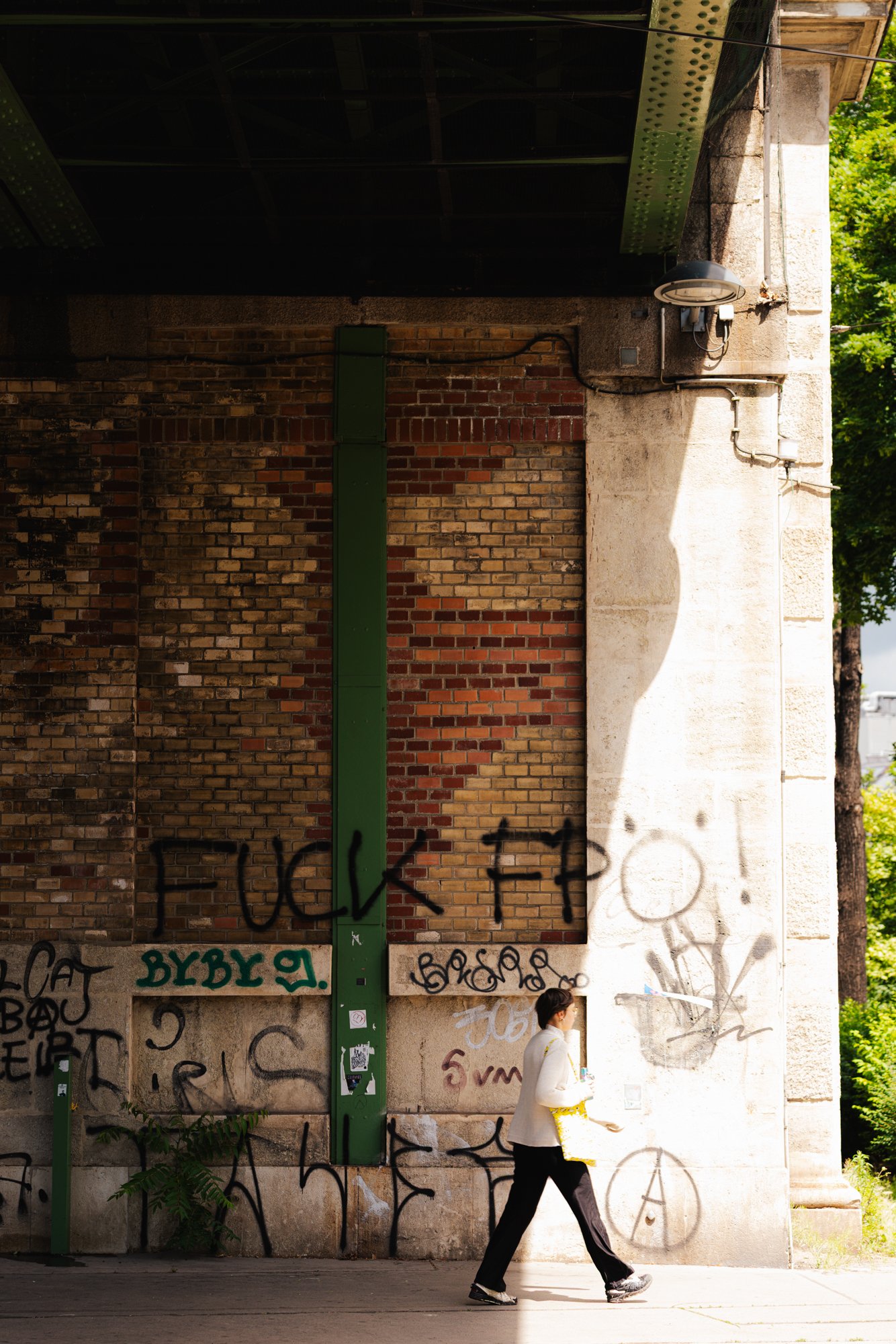 A woman in a white jacket with yellow accents is walking past a wall covered in graffiti. The wall has a mix of brick and concrete with various tags and messages, and there's a green pole in front of it. Bright daylight and green trees are visible on