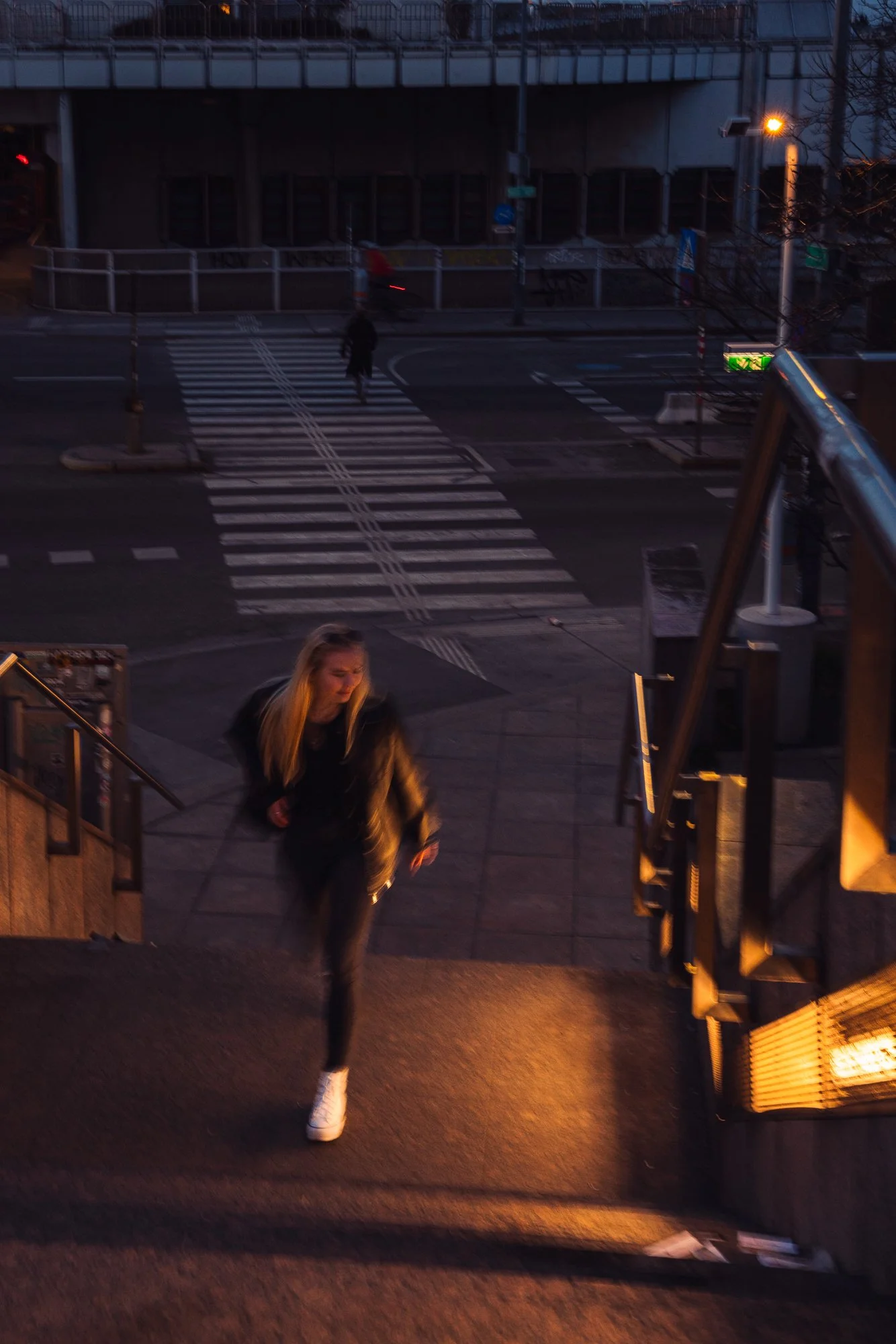 A woman with blonde hair wearing a black jacket and white shoes walking up stairs outside at dusk, with streetlights and a crosswalk visible in the background.