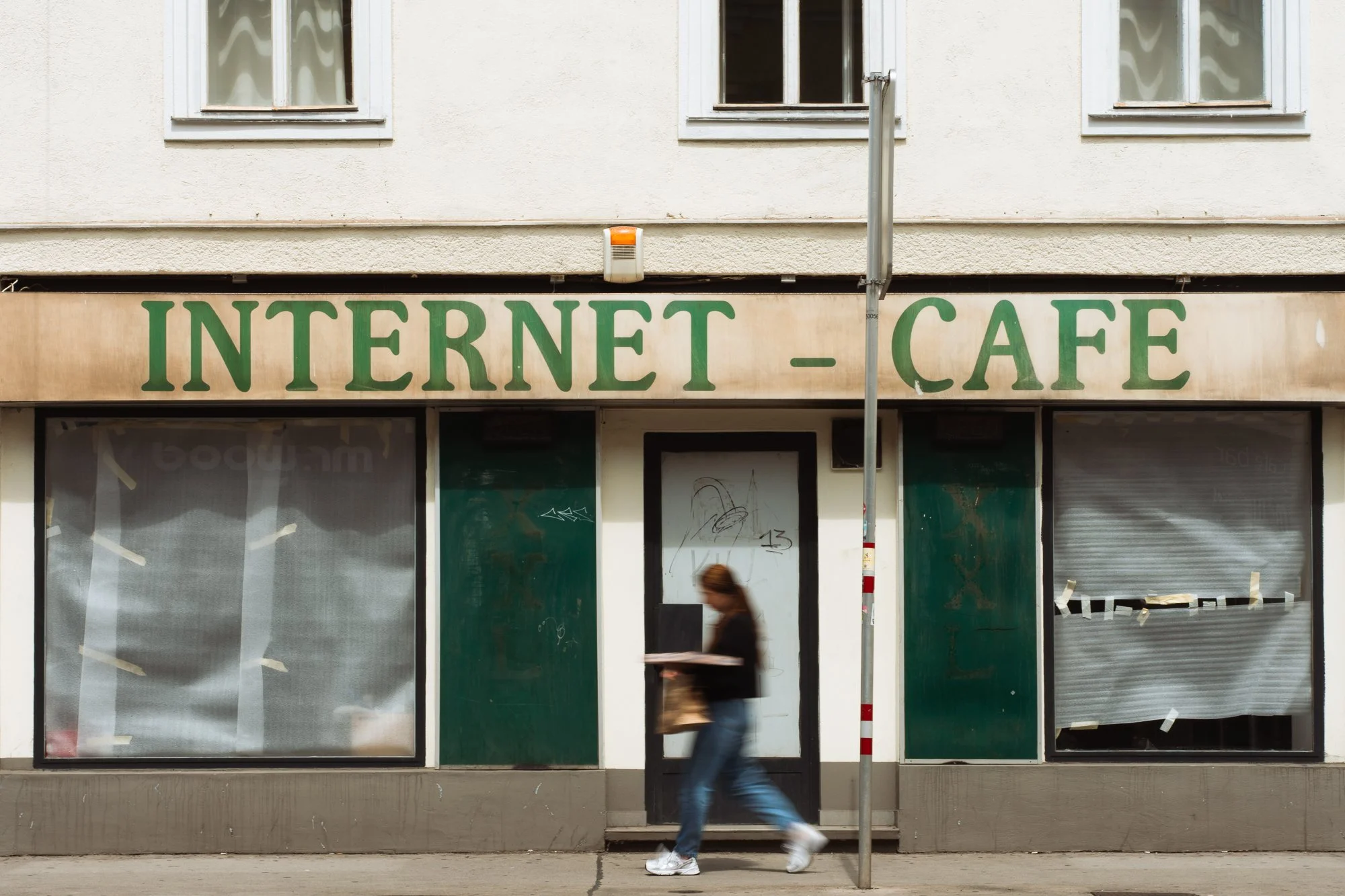 Empty storefront with a sign 'INTERNET CAFE' on top, and a pedestrian walking past in front of it, with a pole in the foreground.