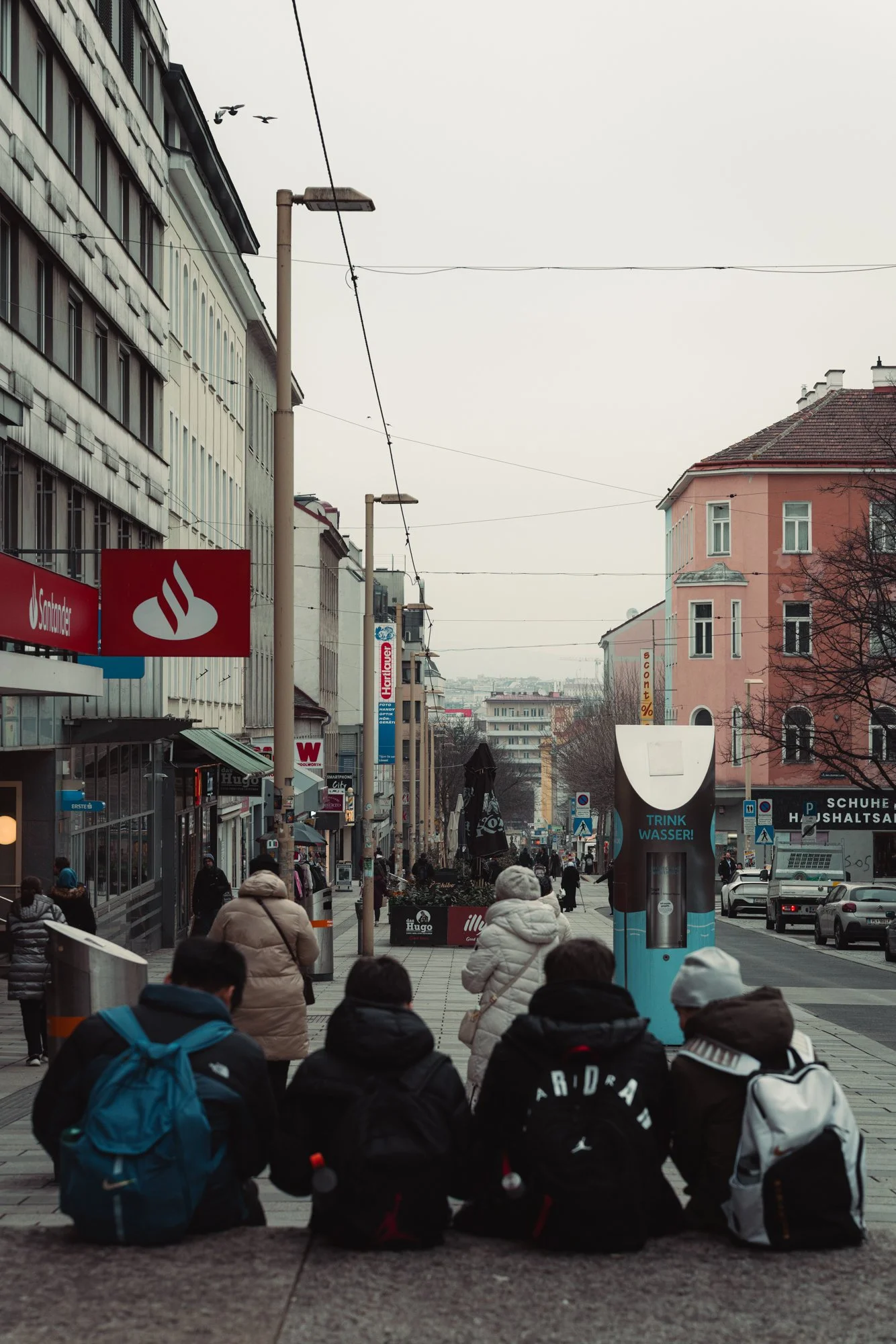 City street scene with pedestrians walking along the sidewalk, some sitting on the ledge, surrounded by buildings with storefronts and advertisements, overcast sky.