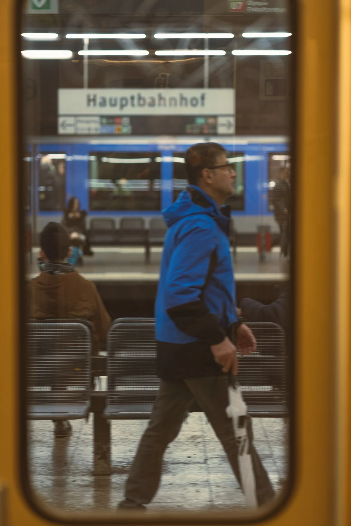 A man wearing glasses and a blue jacket walking through a train station, seen through a glass window with a sign that reads 'Hauptbahnhof' in the background.