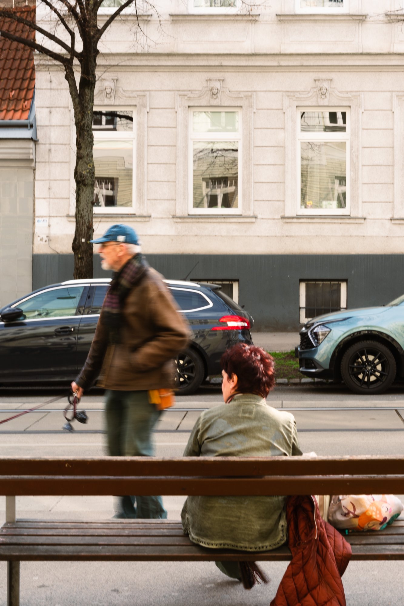 A street scene showing a man walking past cars, two women sitting on a bench, and a building with three windows.