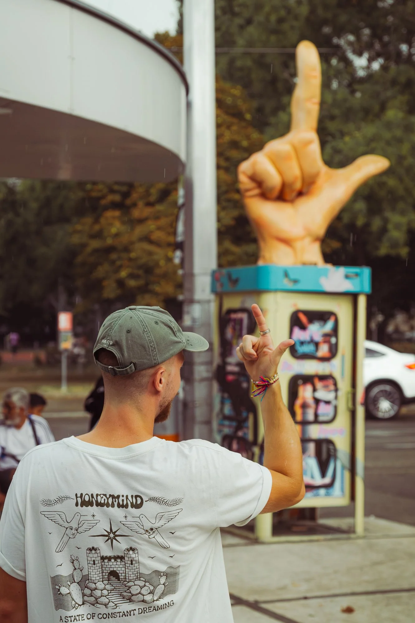 A man wearing a gray baseball cap and a white t-shirt with a tattoo and castle illustration is pointing at a large foam hand sculpture making a shaka sign. The foam hand sculpture is mounted on a yellow cart in an outdoor setting with trees and peopl