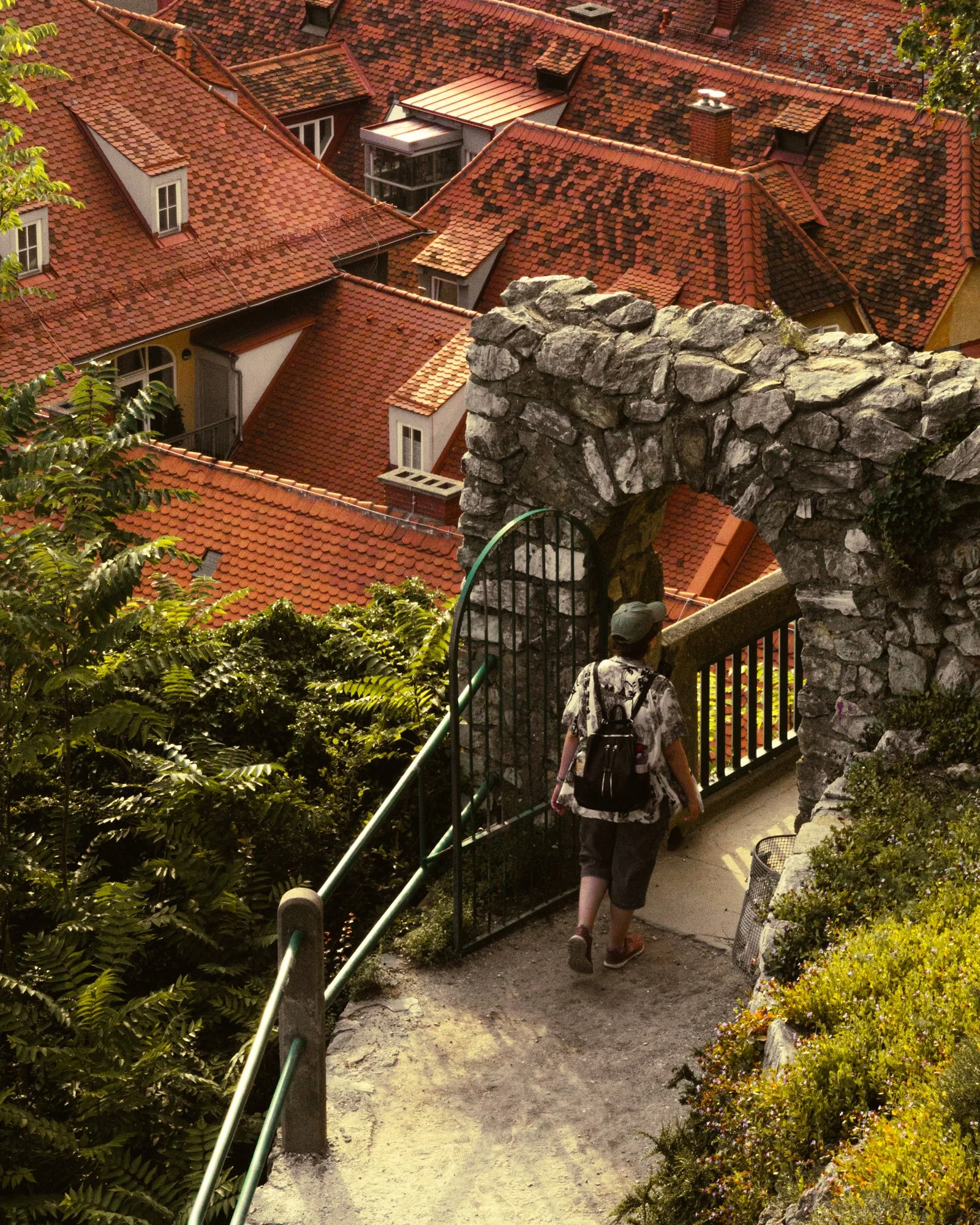 A woman wearing a patterned shirt, carrying a backpack, walking down a stone stairway surrounded by lush greenery. She is descending past a stone archway and a black metal gate, with a hillside view of rooftops with red tiles and dormer windows in th