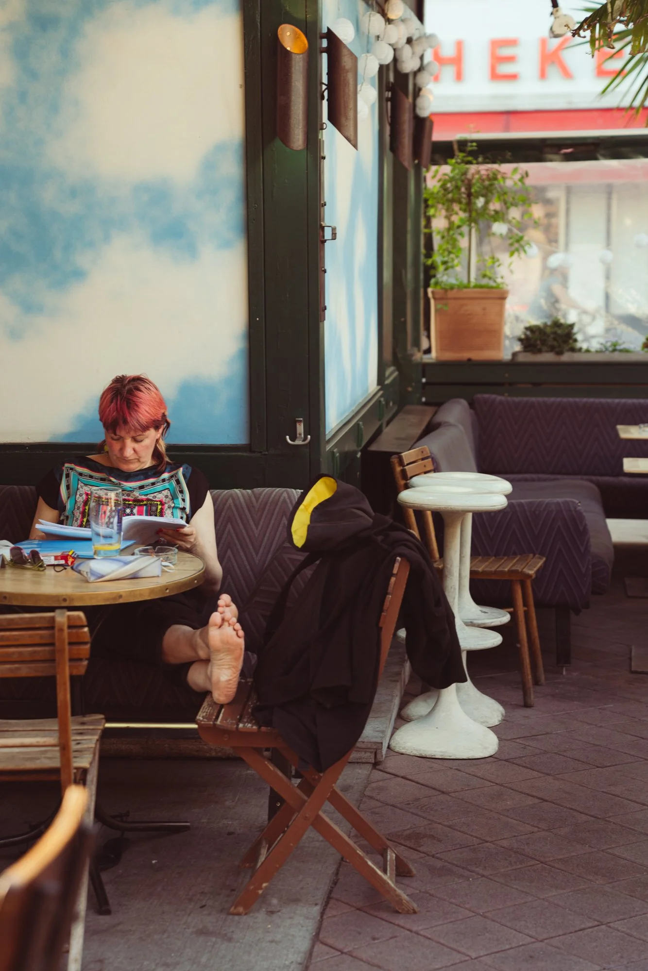 A woman with pink hair reading a menu or a book at an outdoor cafe. She is barefoot, sitting on a wooden chair, with a black jacket draped over her chair. The scene shows a cozy seating area with various chairs and tables, and a blue sky mural on the