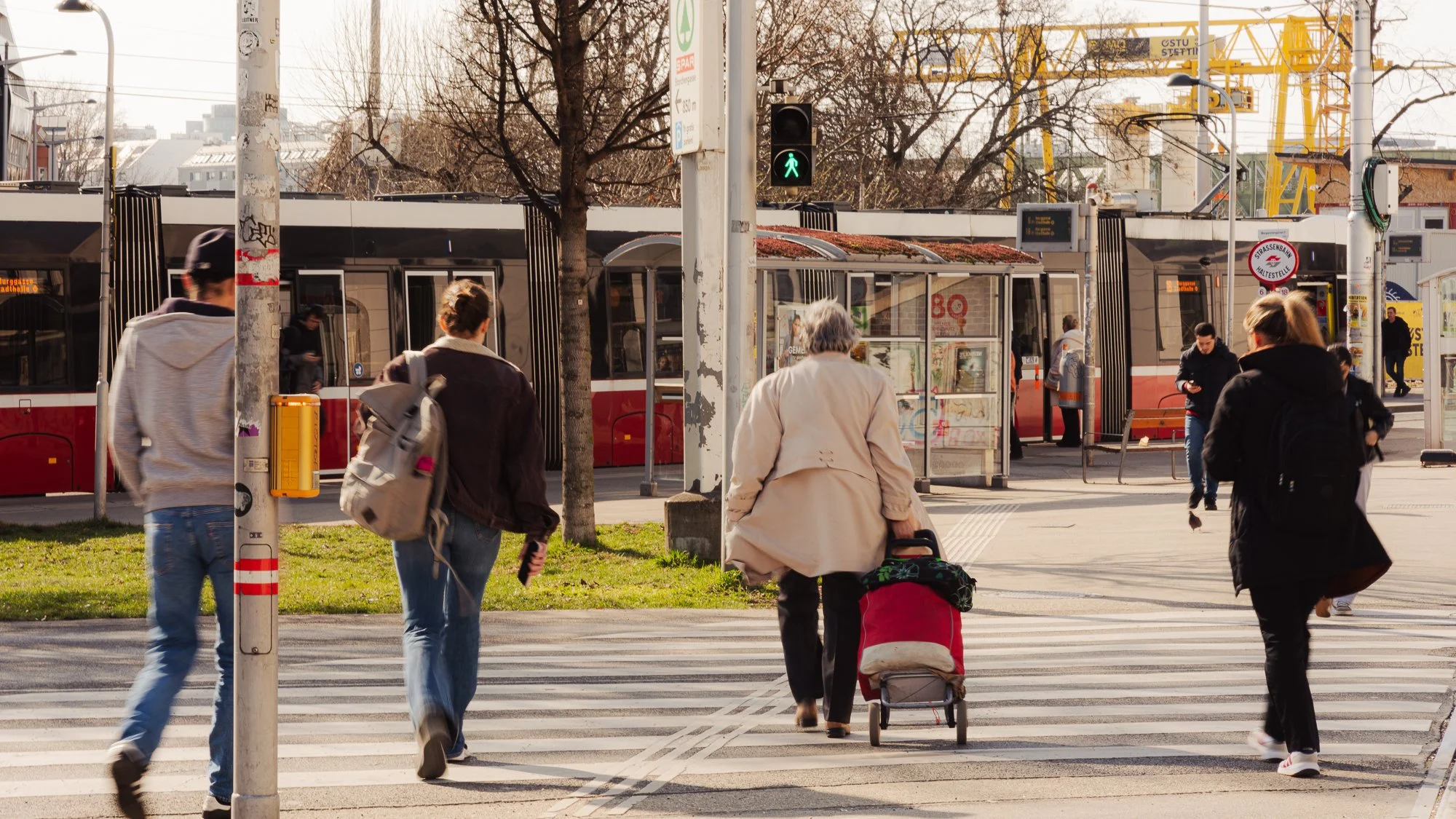 People crossing a street at a crosswalk near a tram stop with a green pedestrian signal