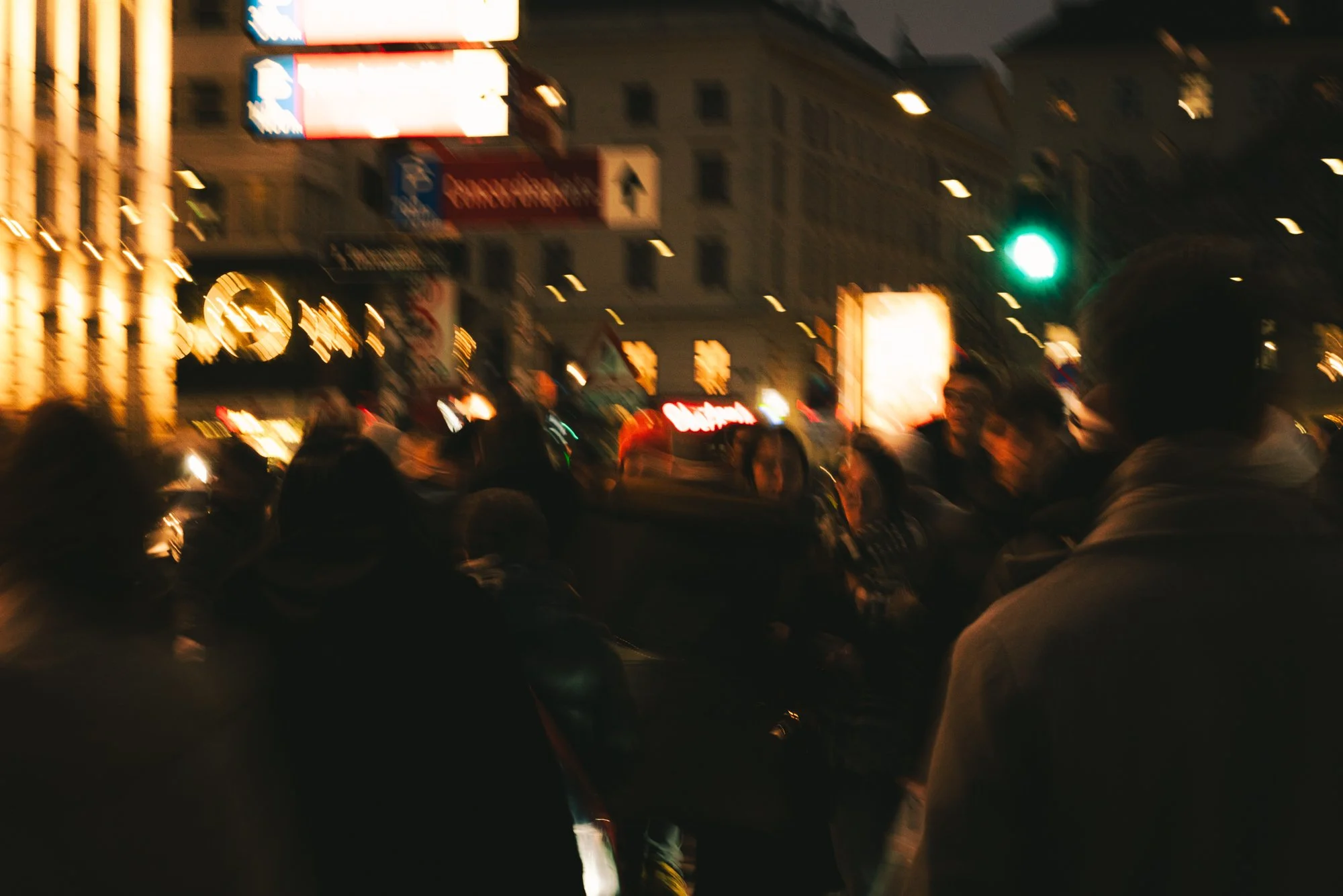 Nighttime city street scene with blurred lights, buildings, and a green traffic light, with a crowd of people in the foreground.
