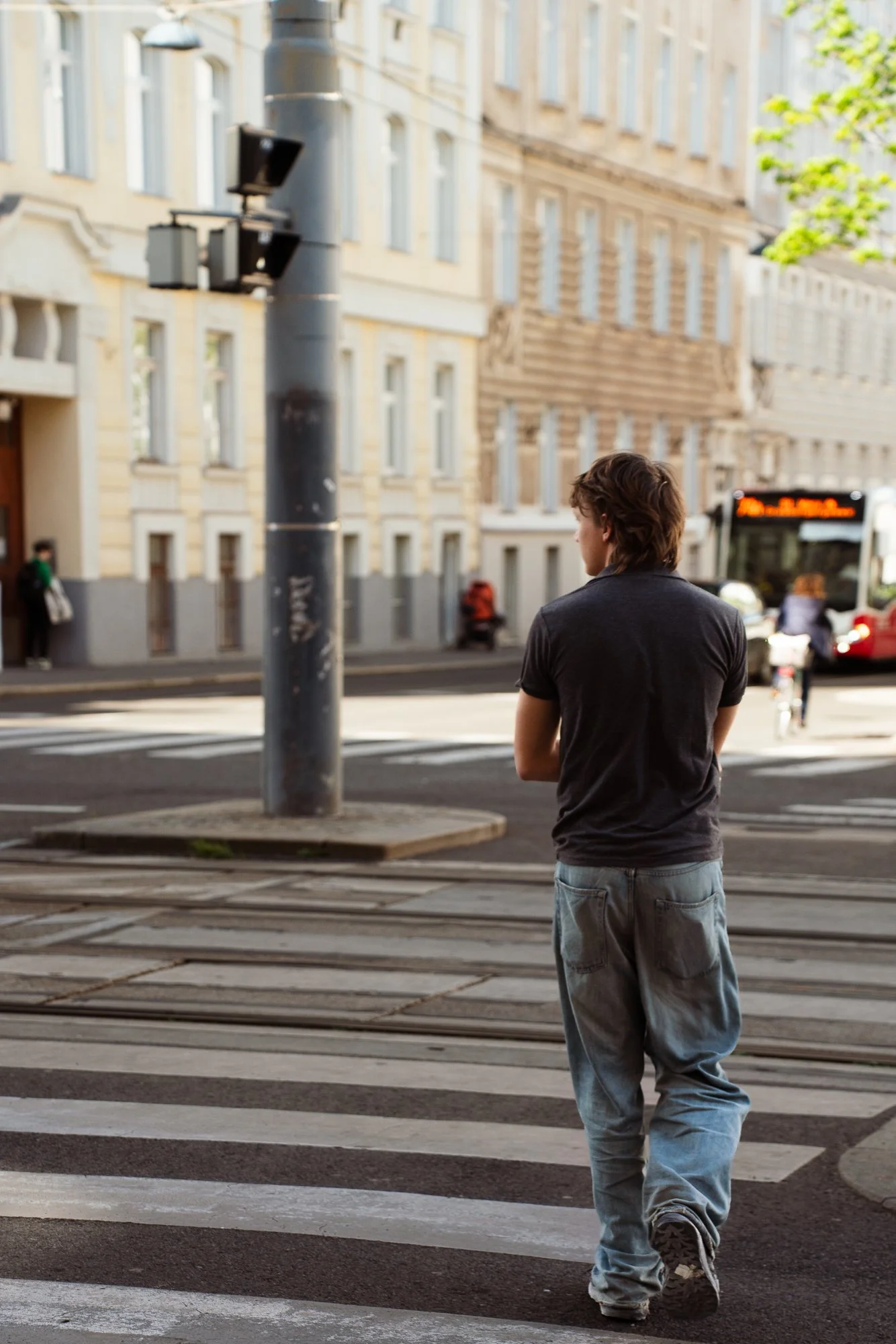 A young man with brown hair, wearing a dark t-shirt and loose jeans, walking across a city street with a crosswalk. There are multi-story buildings, a bus, and other pedestrians in the background.