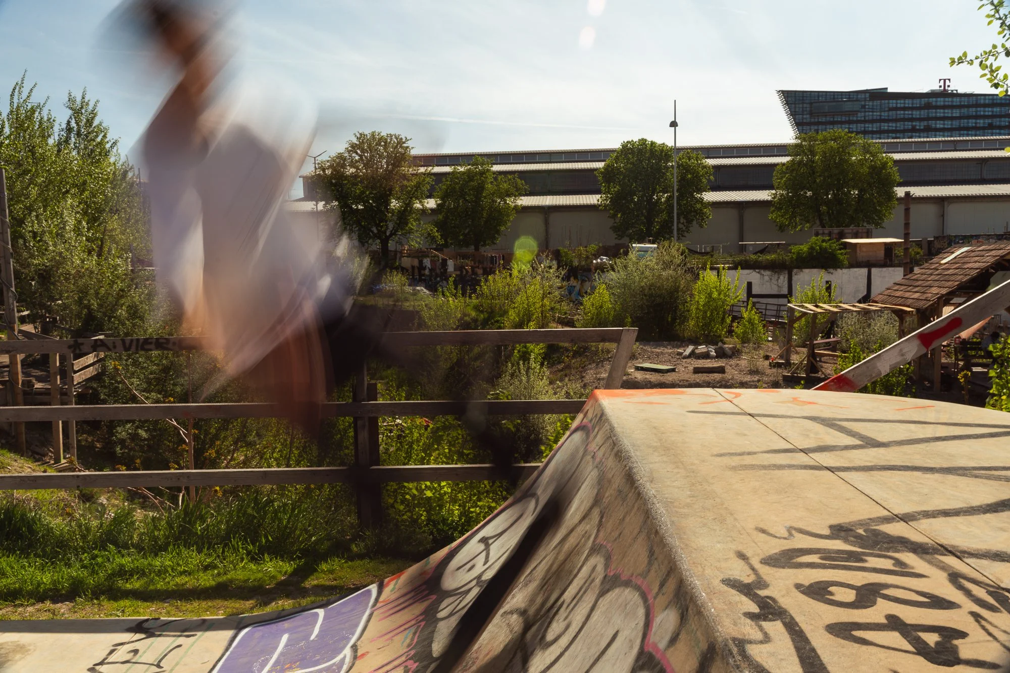 Blurred person in motion on the left, skateboard or skatepark features in the foreground, trees, industrial buildings, and a modern office building in the background on a sunny day