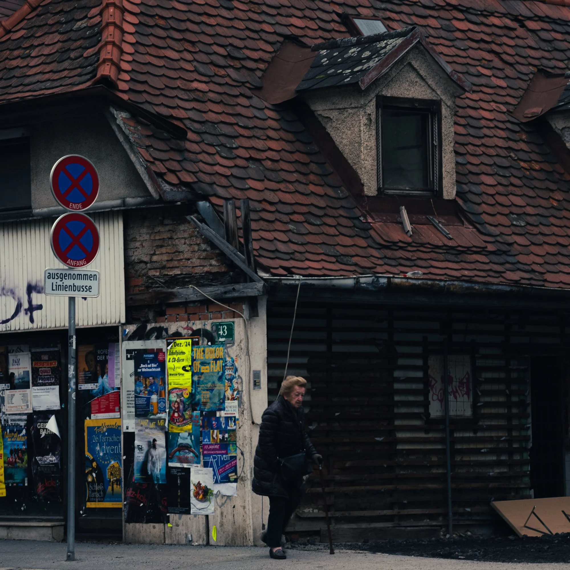 A woman with short hair wearing dark clothing walks with a cane in front of a building with a red tile roof, posters, and signs on the wall, and graffiti on the wall.