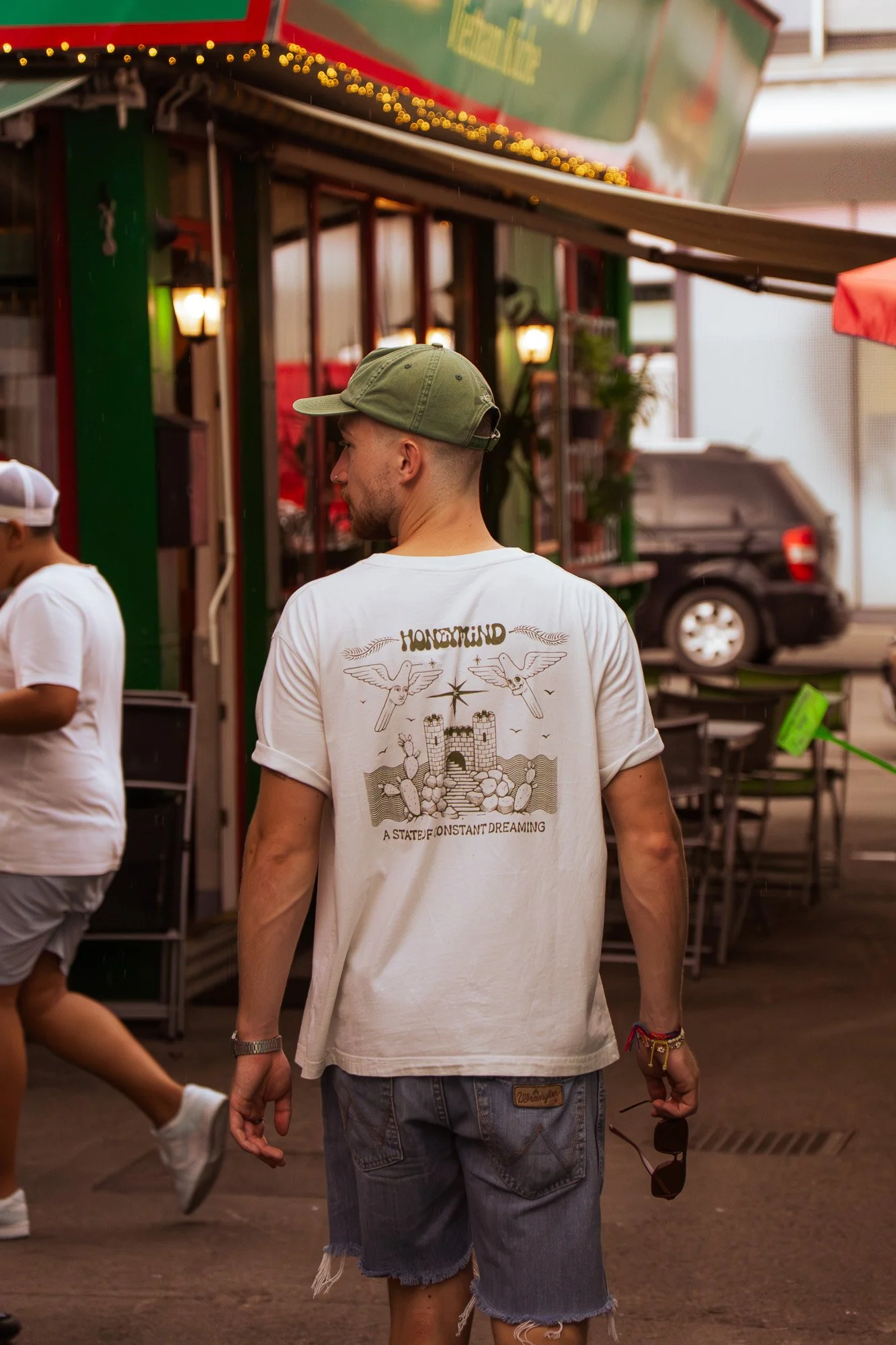 Person wearing a green cap and a white T-shirt with a graphic design, walking in a market or outdoor shopping area, holding sunglasses in their right hand.