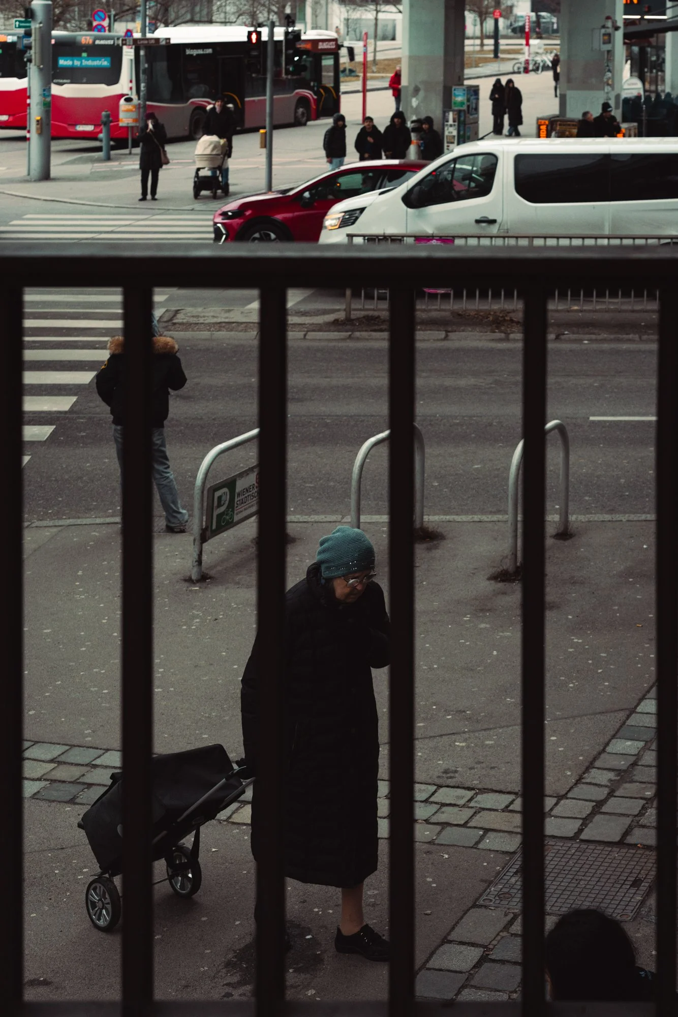 View through a metal railing showing a city street with pedestrians, cars, and a bus, with a woman with a stroller and a woman with a rolling suitcase in the foreground.