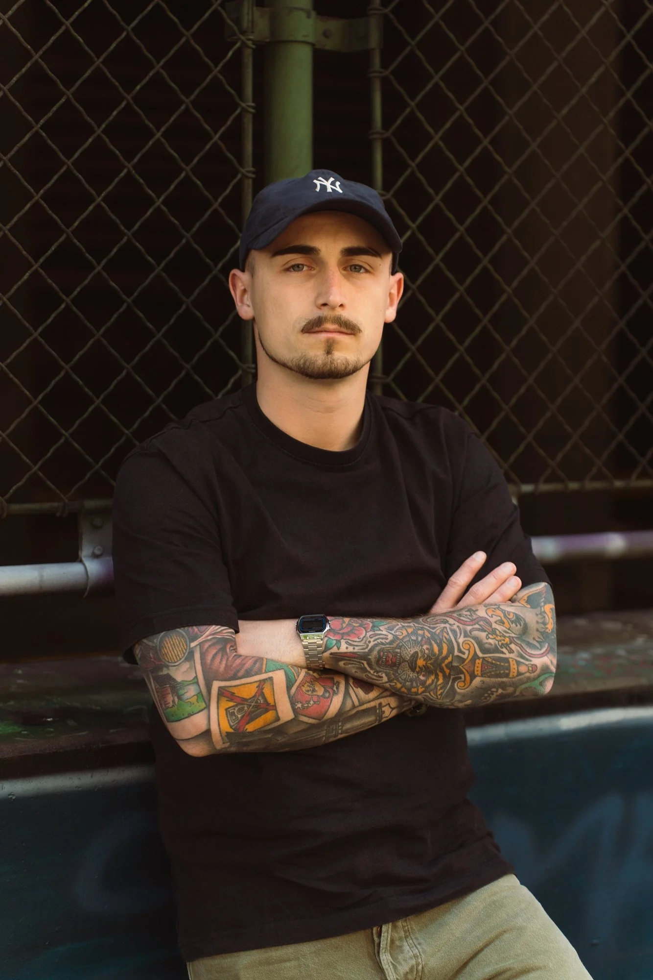 A young man with light skin, short dark hair, a mustache and goatee, wearing a dark baseball cap and black T-shirt, standing in front of a chain-link fence, crossing his arms, showcasing colorful tattoos on both arms, and wearing a silver smartwatch 