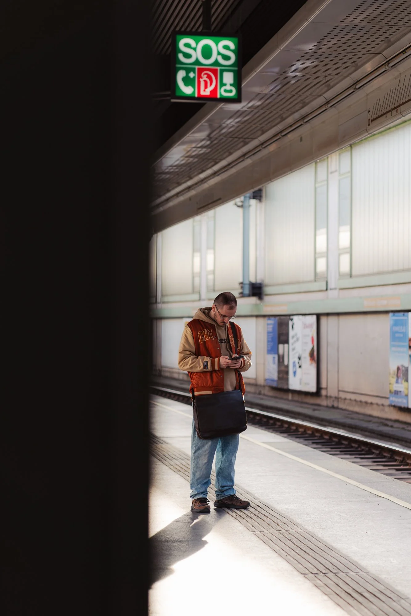 A man stands on a train platform looking at his phone, with a large black object partially obscuring the view, and signage indicating emergency services hanging from the ceiling.