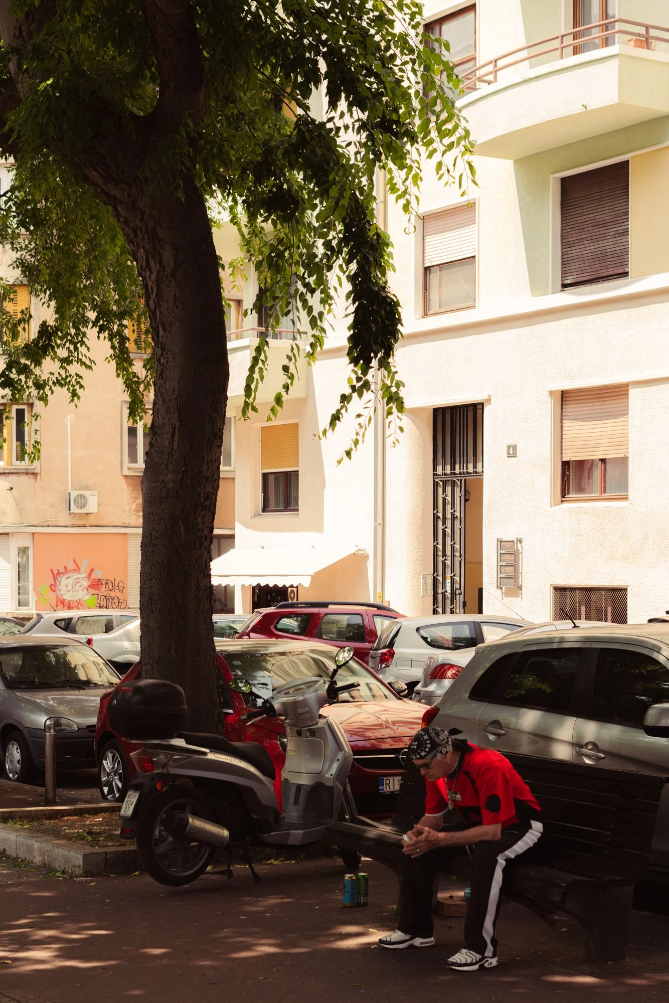 A scene of a parking lot with parked cars and motorcycles under a tree. A person wearing a red jacket and black and white pants is sitting on a bench, looking at their phone, with graffiti visible on a wall in the background.