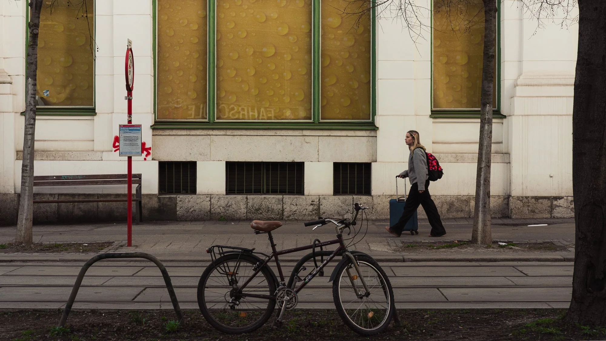 A woman with blond hair wearing glasses, a gray hoodie, and black pants, pulling a blue suitcase while walking a bicycle on the sidewalk in front of a building with large windows and trees.