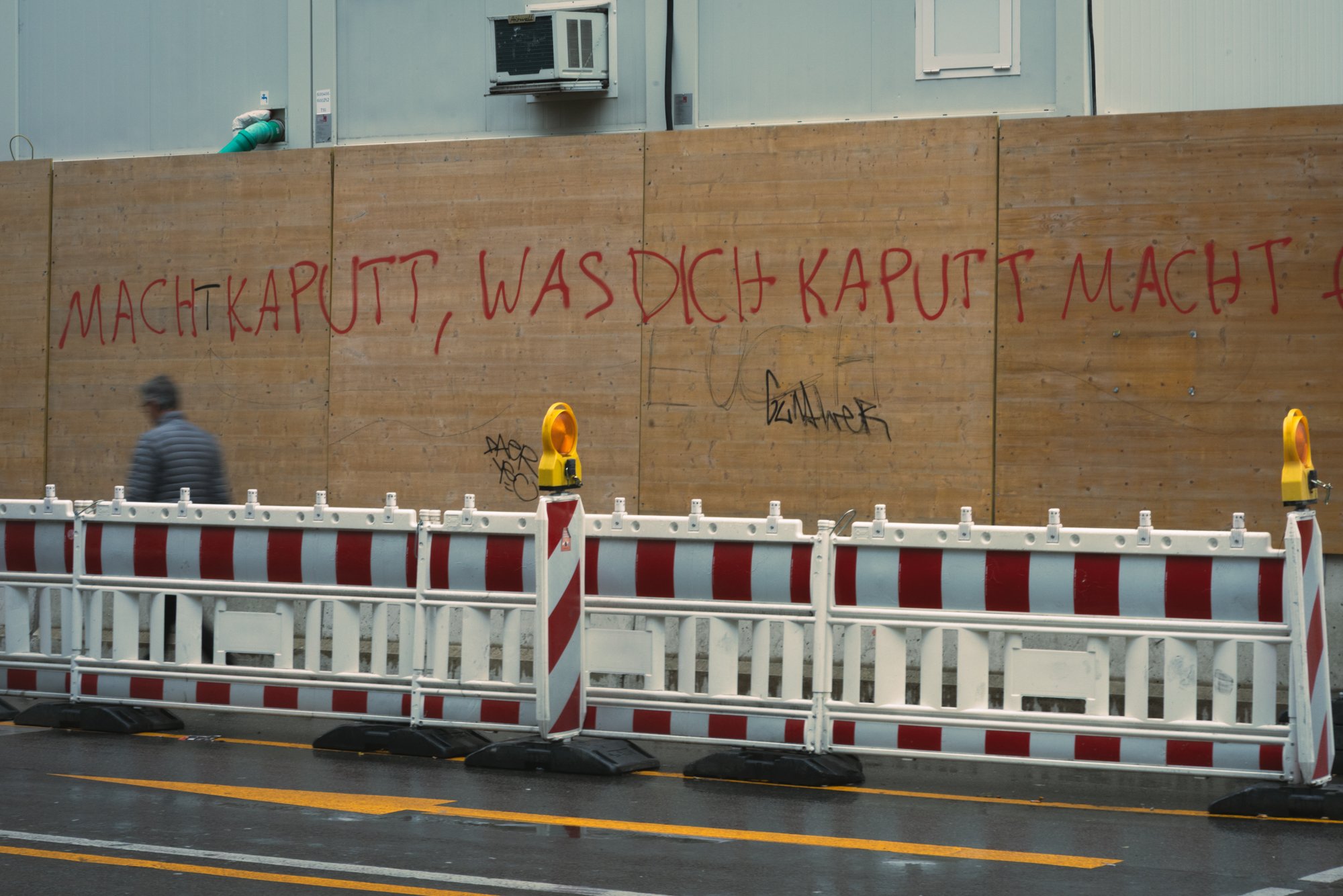A construction site with a wooden safety barrier displaying German graffiti, a person walking past in a striped jacket, and traffic barriers in the foreground on a rainy day.