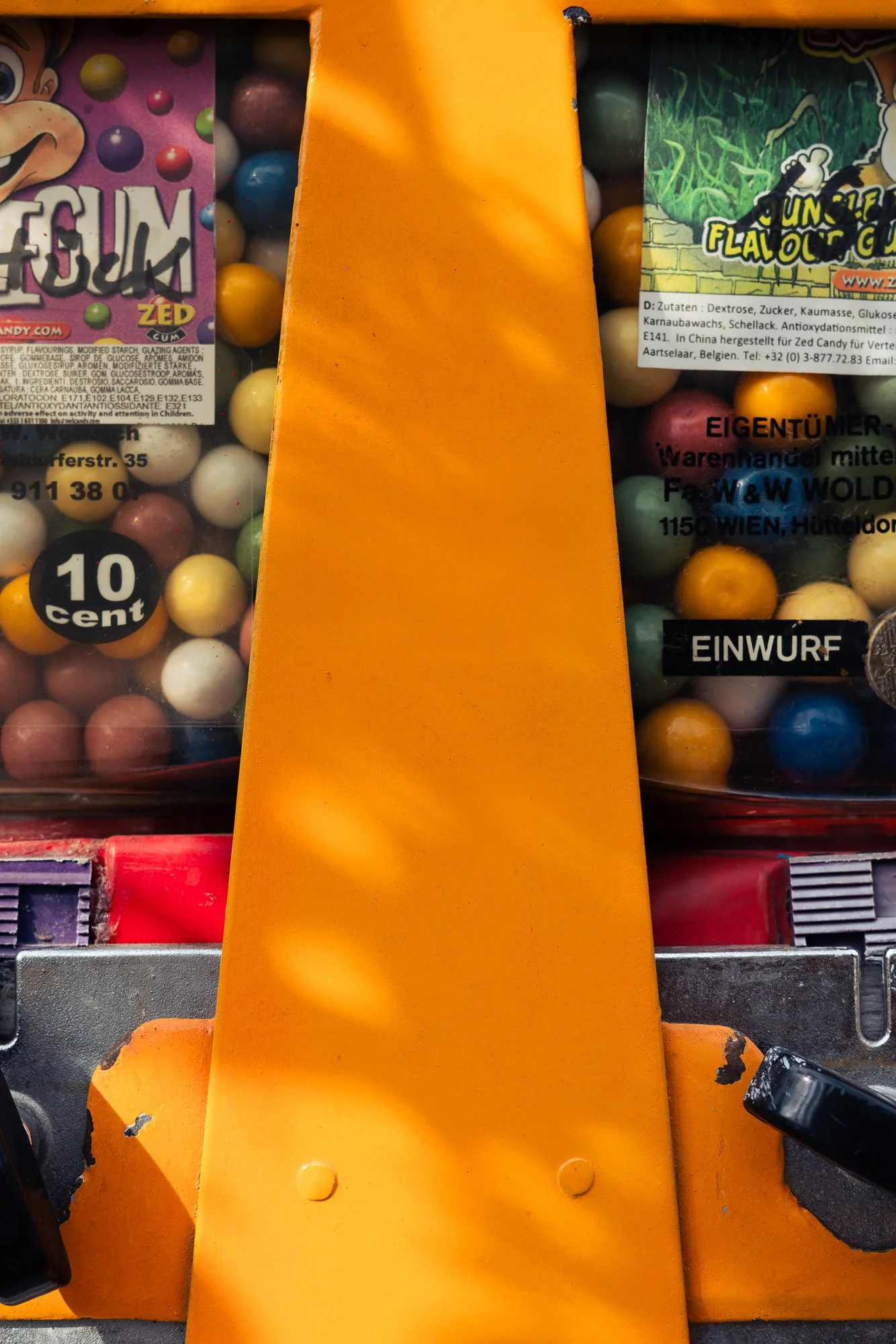 Close-up of a colorful arcade or pinball machine with a yellow-orange plastic ramp and multicolored gumballs behind a glass window, with some text labels visible.