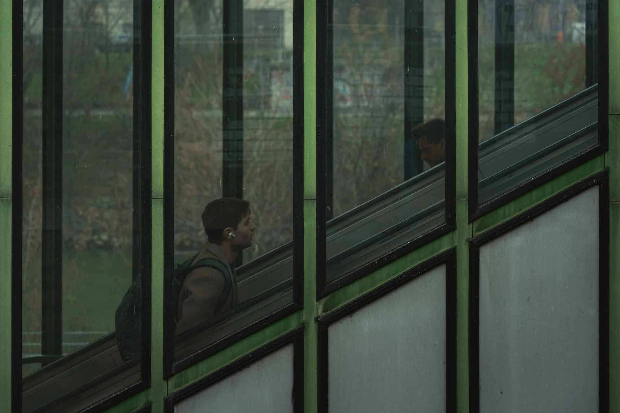 A young man walking up a staircase inside a building, seen through large glass windows with black frames and green accents.